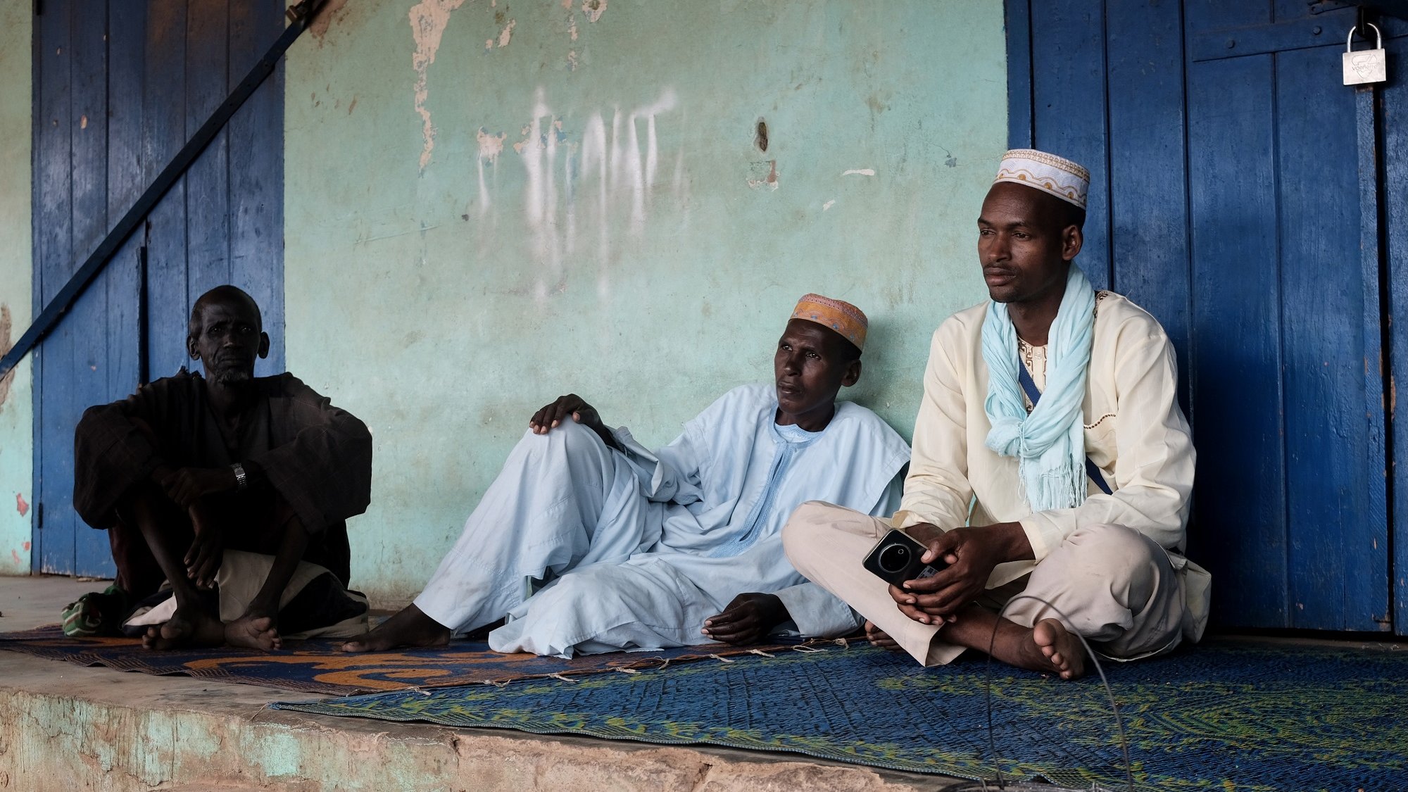 Cette image montre trois hommes assis sur un tapis, à l'extérieur d'un bâtiment aux murs de couleur turquoise. L'atmosphère semble calme et détendue. Ils portent des vêtements traditionnels : deux d'entre eux sont vêtus de tuniques, tandis que le troisième porte un chapeau. L'un des hommes est assis en tailleur, tandis que les deux autres sont en position plus détendue, le dos appuyé contre le mur. À côté d'eux, il y a un petit seau en métal d'où s'écoule un peu d'eau. En arrière-plan, des murs sont partiellement recouverts de graffiti. L'environnement semble rural, avec une lumière douce qui illumine la scène.
