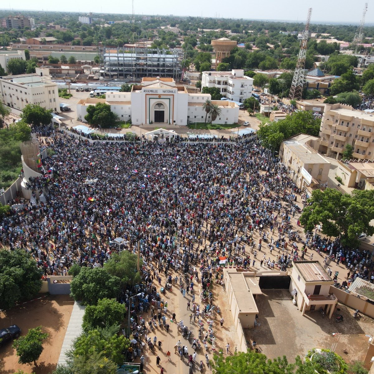 L'image montre une grande foule rassemblée dans une place publique, probablement lors d'une manifestation ou d'un événement collectif. Les personnes sont regroupées en masse, et on peut imaginer une ambiance vibrante, avec des slogans ou des chants. En arrière-plan, des bâtiments se dressent, notamment un bâtiment avec une architecture marquée. Les arbres entourent la zone, ajoutant une touche de verdure. L'atmosphère semble dynamique, illustrant l'engagement de la communauté dans une cause.