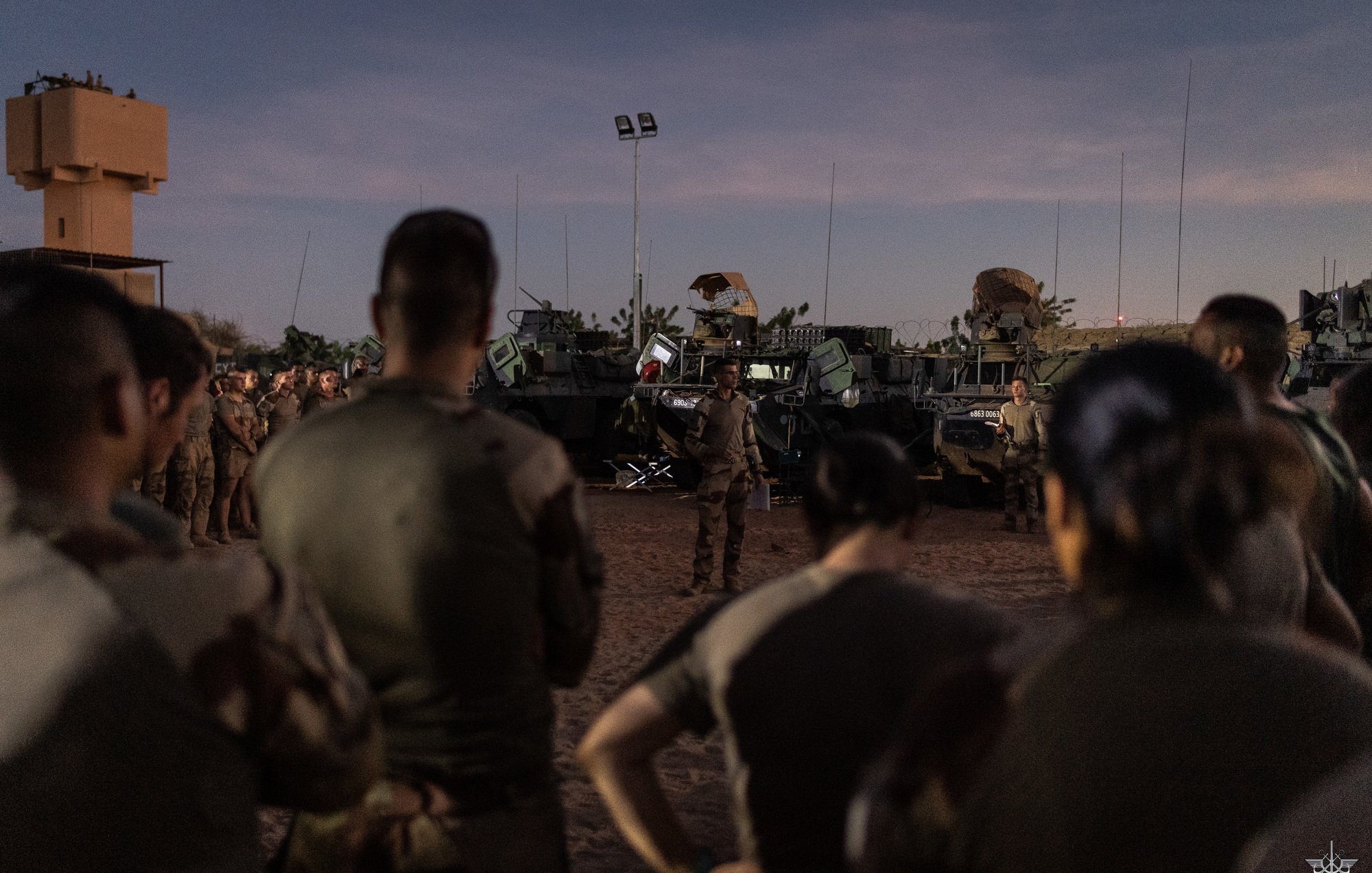 L'image montre un groupe de soldats rassemblés, attentifs, dans un environnement désertique au crépuscule. Ils sont partiellement de dos, ce qui met en avant leur tenue militaire. À l'arrière-plan, plusieurs véhicules militaires sont visibles, équipés d'armements. Le ciel est teinté de nuances douces, marquant la transition entre le jour et la nuit. L'atmosphère semble concentrée, suggérant une préparation ou un briefing important pour les soldats sur le terrain.
