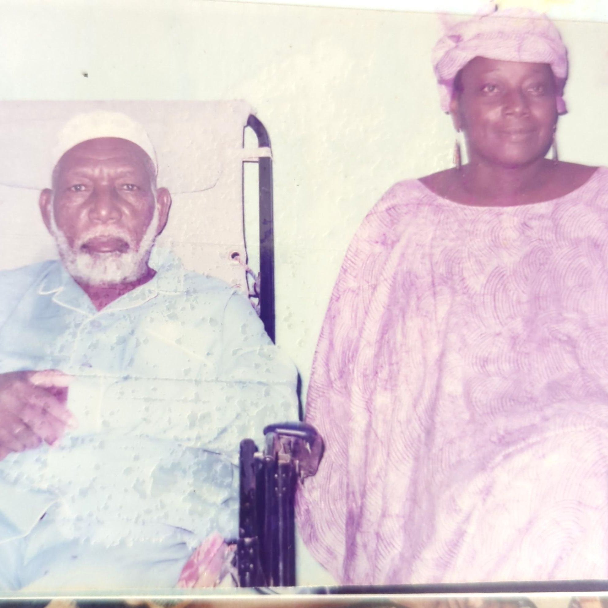 L'image montre un homme et une femme assis côte à côte. L'homme, plus âgé, est en fauteuil roulant et porte une tenue traditionnelle, comprenant un turban blanc. Il a une barbe grise et semble contemplatif. À côté de lui, la femme, qui a également des traits traditionnels, est vêtue d'une robe fluide et d'un foulard. Elle a une expression douce et chaleureuse. Le fond de l'image est sobre, créant un contraste avec leurs vêtements. L'atmosphère dégage une sensation de respect et de sérénité.