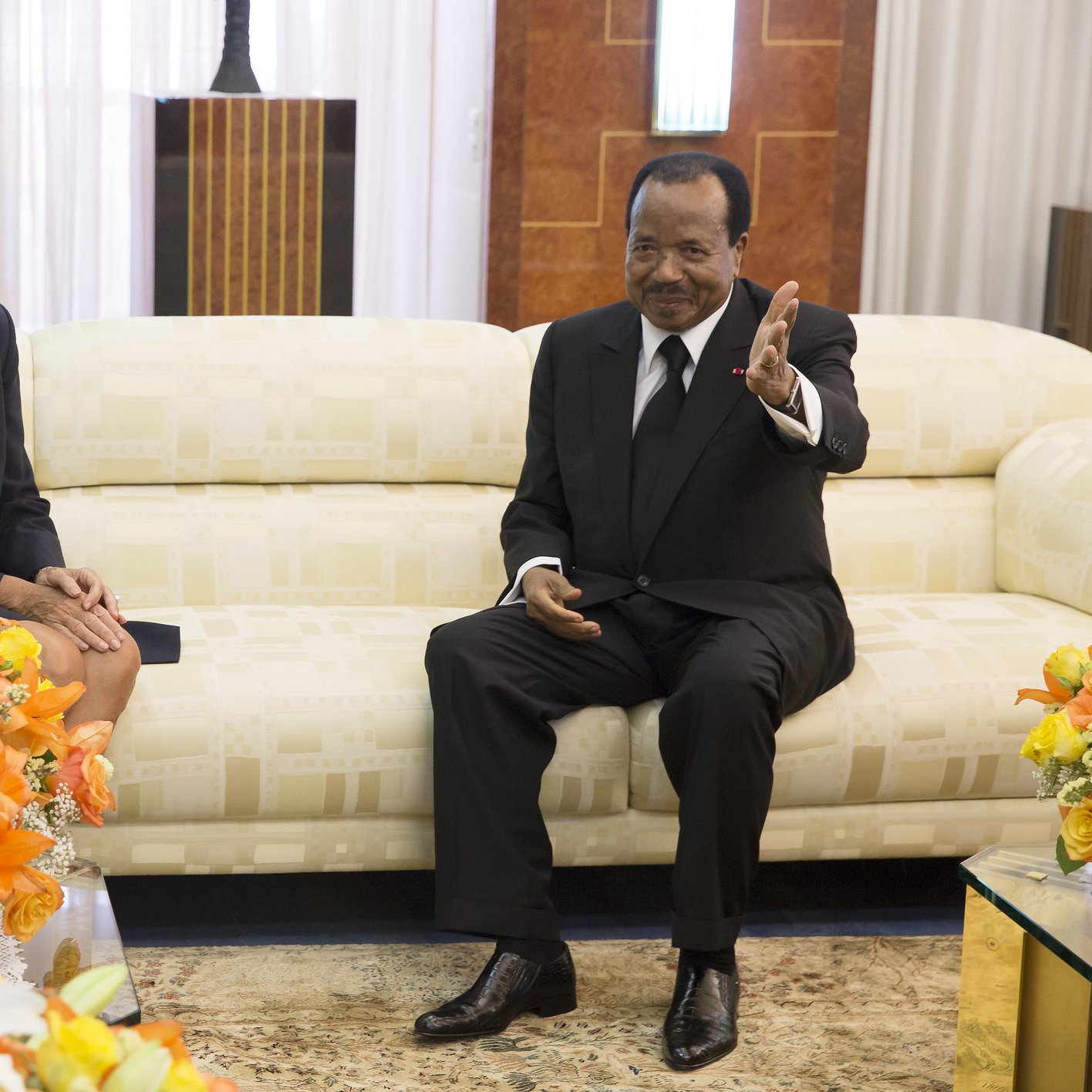 L'image montre deux personnes assises dans un salon élégant. À gauche, une femme aux cheveux courts et blonds, portant un blazer noir avec une écharpe rouge, sourit et semble détendue. À droite, un homme en costume noir et chemise blanche, assis sur un canapé crème, lève la main en signe de bienvenue ou pour parler. Ils sont entourés de bouquets de fleurs colorées, dominés par des teintes orange et jaune, qui ajoutent une touche de chaleur à l'environnement. Le décor est raffiné, avec des murs en bois et un éclairage doux, créant une ambiance conviviale.