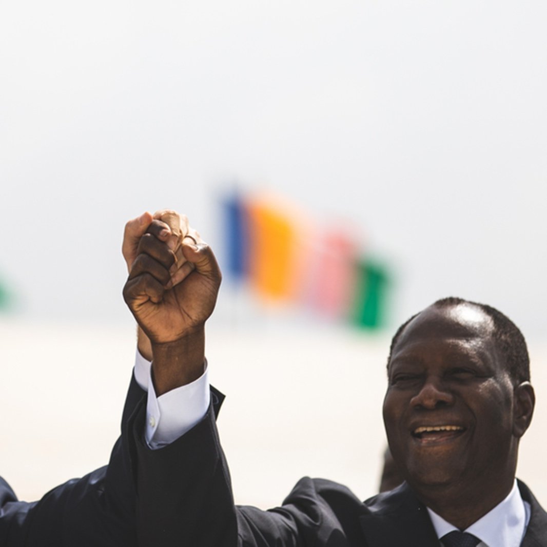 Imaginez une scène marquée par un moment de camaraderie et d'unité. Deux hommes se tiennent côte à côte, levant la main droite en l'air, leurs poings serrés, symbolisant la force et la solidarité. À gauche, on perçoit un homme avec une coupe de cheveux soignée, vêtu d'un costume sombre, tandis que l'homme à droite, également en costume, affiche un grand sourire chaleureux. En arrière-plan, plusieurs drapeaux flottent doucement au vent, représentant différentes nations. L'atmosphère est festive et pleine d'espoir.