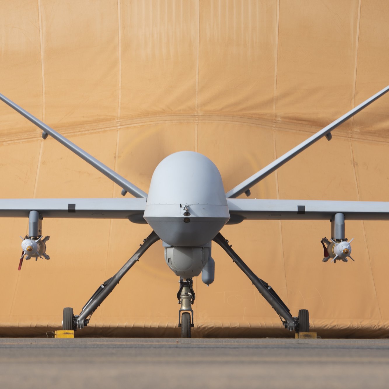 L'image montre un drone militaire de type MQ-9 Reaper. Il est positionné de face, avec le corps fuselé et l'aile légèrement étendue. À chaque côté du fuselage, on peut voir des réservoirs ou des munitions. En arrière-plan, il y a une toile beige qui semble un hangar ou une protection, lui donnant un aspect de préparation à un vol. Le sol est en béton, créant un environnement d'aérodrome. L'ensemble est impressionnant, évoquant la technologie aéronautique moderne.