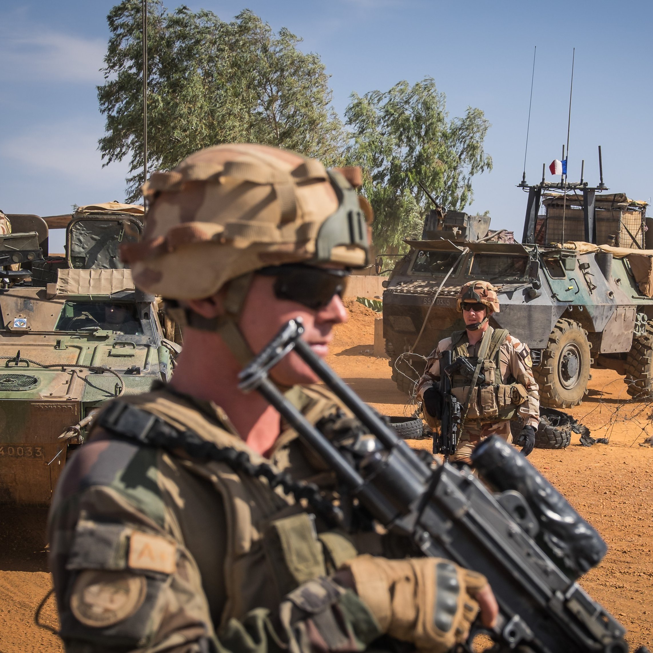 The image depicts a military scene in a desert environment. In the foreground, a soldier wearing a helmet and sunglasses is holding an assault rifle, looking serious and vigilant. Behind him, there are other soldiers and military vehicles, including an armored personnel carrier. The background shows a clear blue sky with some greenery and sand, indicating a remote and arid location. The overall atmosphere suggests a sense of readiness and military operations.