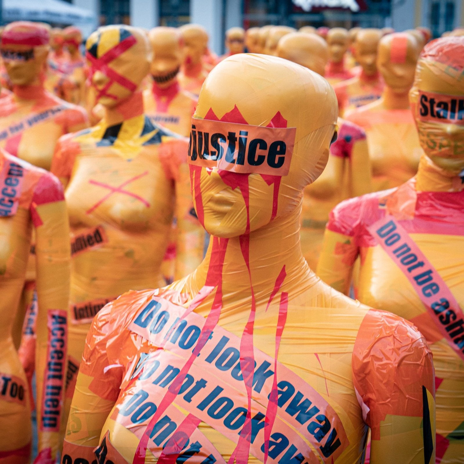 L'image montre une installation artistique composée de nombreux mannequins recouverts de bandes de plastique colorées, principalement dans des teintes orange et jaune. Ces mannequins sont alignés et se font face, créant une impression de masse. Sur leur surface, on peut voir des mots et des phrases comme "justice", "Stalking", "MeToo" et "Do not look away", écrits en noir et rouge, ce qui suggère un message fort et engagé. L'ambiance générale semble à la fois frappante et troublante, évoquant des thèmes de lutte contre l'injustice et les violences faites aux femmes. Les couleurs vives et les slogans bien en vue attirent l'attention et suscitent réflexion.