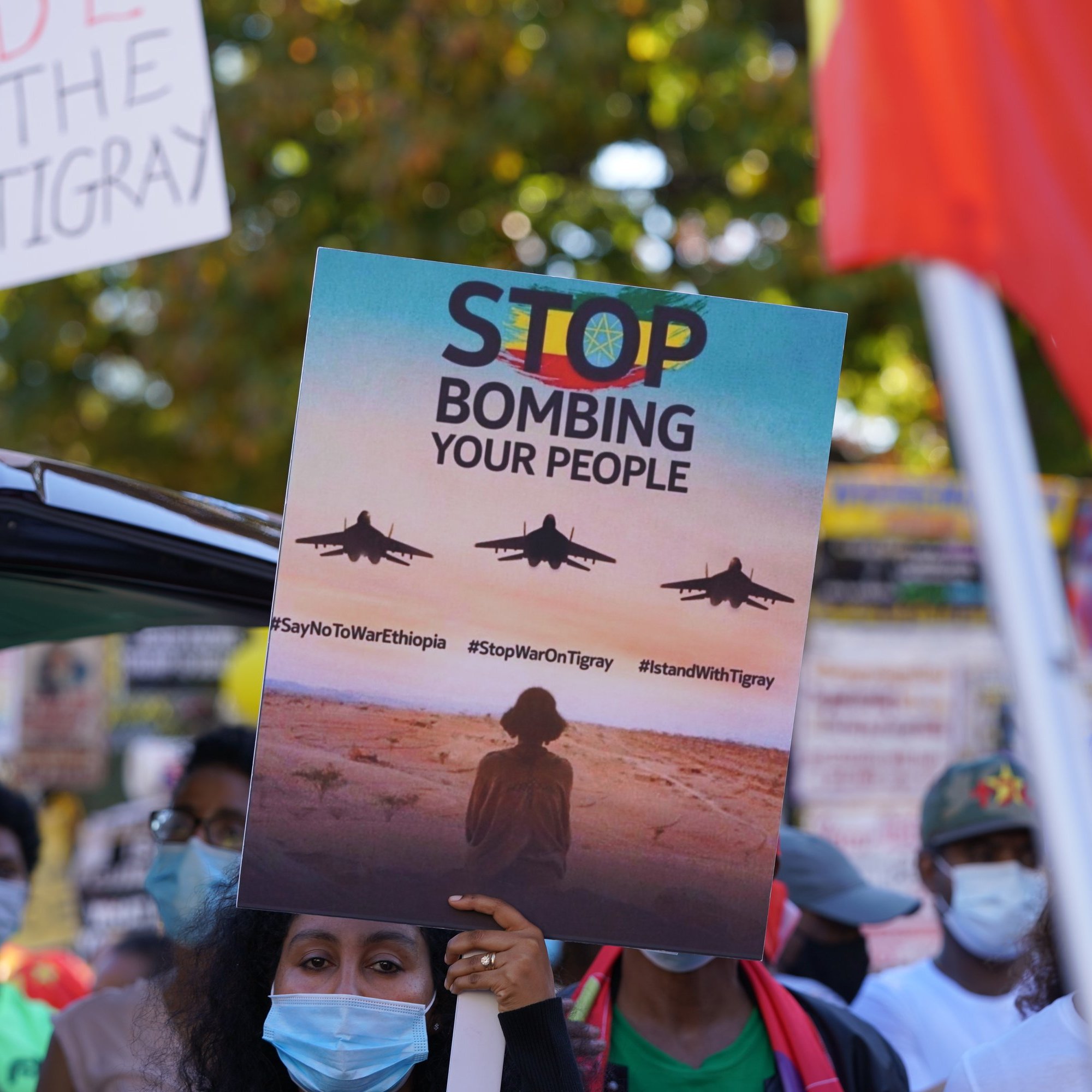 L'image montre une scène de manifestation. Au premier plan, une personne tient une grande pancarte qui affiche le message "STOP BOMBING YOUR PEOPLE" avec des images d'avions de chasse en vol. La personne est entourée d'autres manifestants portant des masques faciaux, certains brandissant d'autres pancartes. En arrière-plan, on peut voir des drapeaux et des affiches qui expriment des messages de protestation. L'atmosphère est chargée d'émotion, soulignant un appel urgent à l'action.