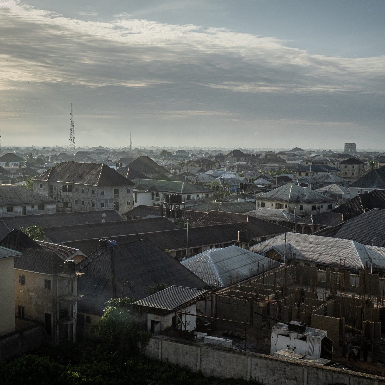 Dans cette image, on voit un paysage urbain d'une ville d'Afrique, probablement au lever du soleil. Les toits de différentes maisons se chevauchent, formant une mosaïque de formes et de textures. La majorité des toits sont en tôle, certains ayant des angles abrupts, tandis que d'autres sont plats. En arrière-plan, on aperçoit des constructions en cours, avec des structures en béton qui indiquent un développement urbain. Le ciel est parsemé de nuages, offrant une lumière douce et diffuse. On devine une atmosphère paisible au matin, avec des ombres légères qui accentuent les contours des bâtiments. Au loin, on distingue des antennes et des colonnes, signes de l'urbanisation moderne. L'ensemble dégage une impression de vie et d'activité, mais aussi de tranquillité à cette heure matinale.