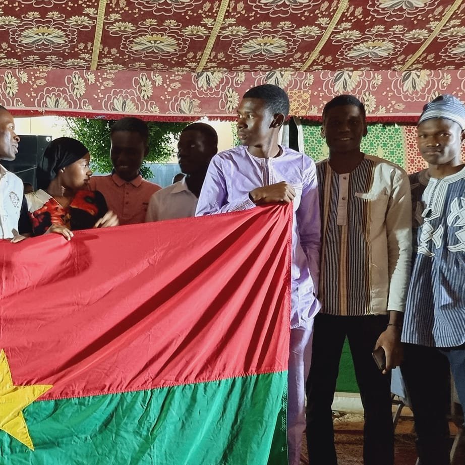 The image depicts a group of young people gathered together, holding a large flag of Burkina Faso, which features a red field with a green stripe at the bottom and a yellow star in the center. The setting appears to be festive, possibly during a cultural or community celebration, with decorative elements in the background. The individuals are wearing a mix of traditional and modern clothing, smiling and posing together, suggesting a sense of unity and pride in their national identity.