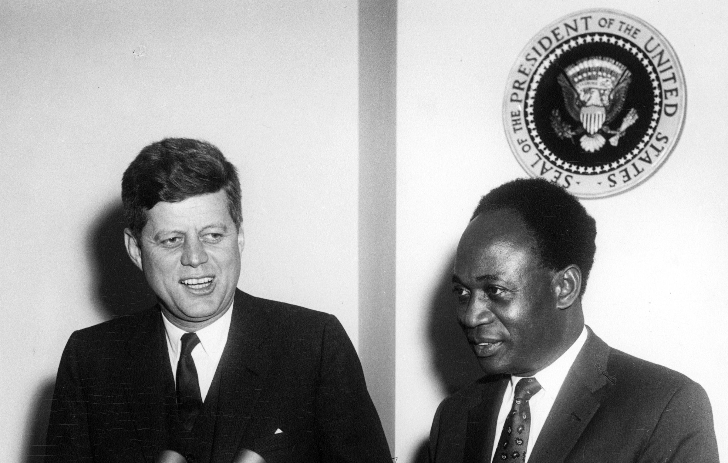 The image features two men, one of whom is John F. Kennedy, the President of the United States, and the other is likely Kwame Nkrumah, the first President of Ghana. They appear to be at a press conference or a formal meeting, with microphones in front of them. Behind them, there is a seal that represents the office of the President of the United States. Both men are dressed formally, with Kennedy in a suit and tie, and they are smiling, suggesting a cordial atmosphere.