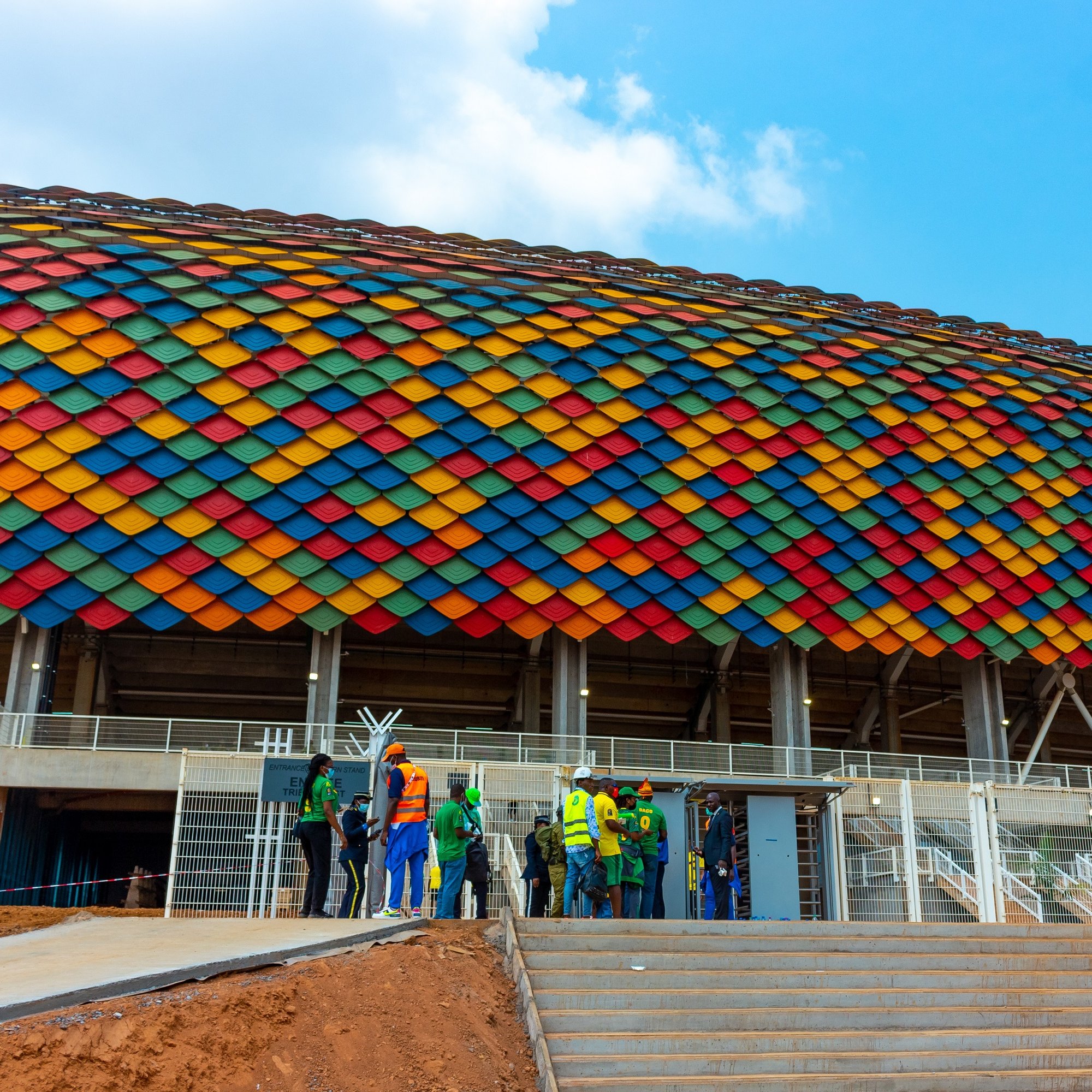 L'image montre un grand stade avec une architecture moderne. La façade du stade est ornée de tuiles de diverses couleurs vives, allant du rouge, orange, jaune, vert, bleu jusqu'au violet, formant une belle mosaïque sur un fond en béton. Au premier plan, on peut voir un groupe de personnes, habillées en vêtements de travail, qui semblent discuter ou se préparer à entrer dans le stade. Les escaliers mènent à une entrée principale, et le ciel au-dessus est bleu avec quelques nuages épars, ajoutant une impression de clarté et d'énergie à la scène.