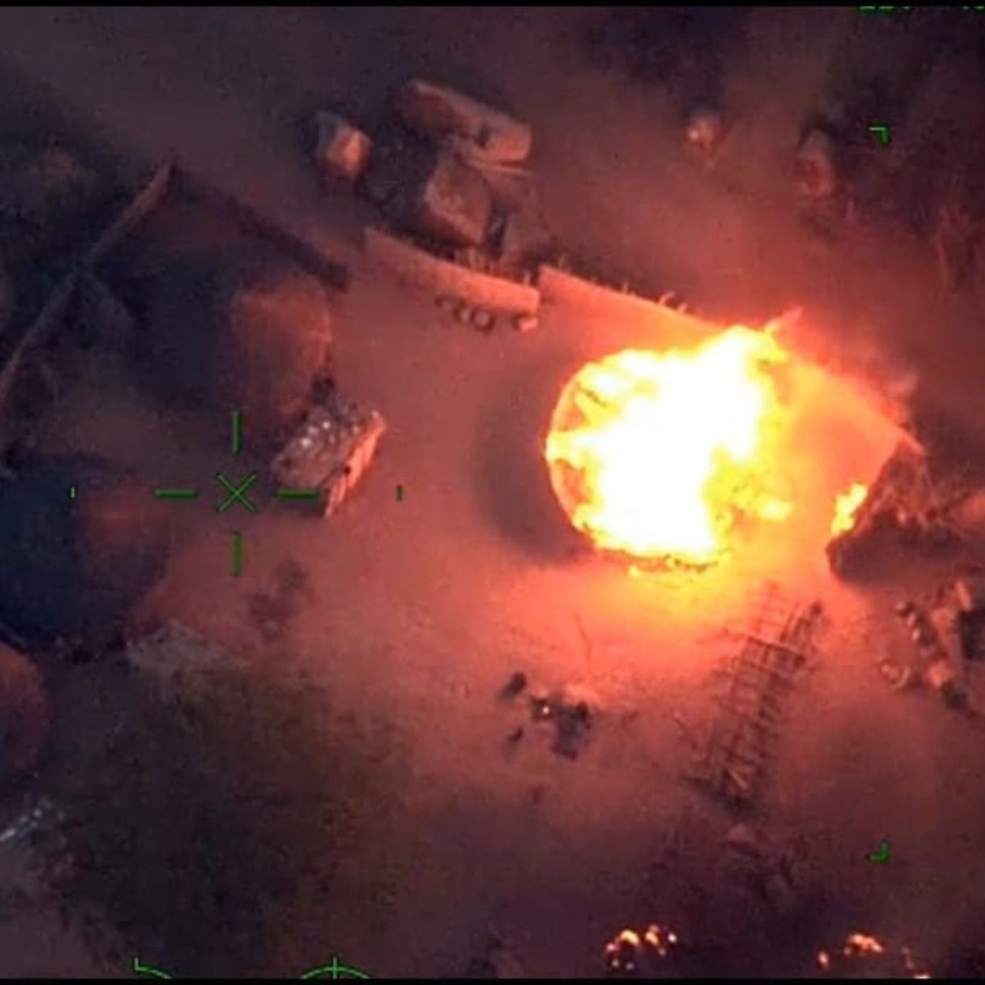 L'image représente une scène vue du ciel, où l'on aperçoit un environnement rural. Au centre, une grande flamme intense monte vers le ciel, créant une illumination vive dans l'obscurité. Entourant cette flamme, on peut distinguer plusieurs structures, probablement des huttes ou des abris, formées de matériaux naturels. Le sol semble légèrement chaotique, avec des débris dispersés et des ombres créées par les objets environnants. L'atmosphère est dramatique et évoque une situation de tension ou de conflit.