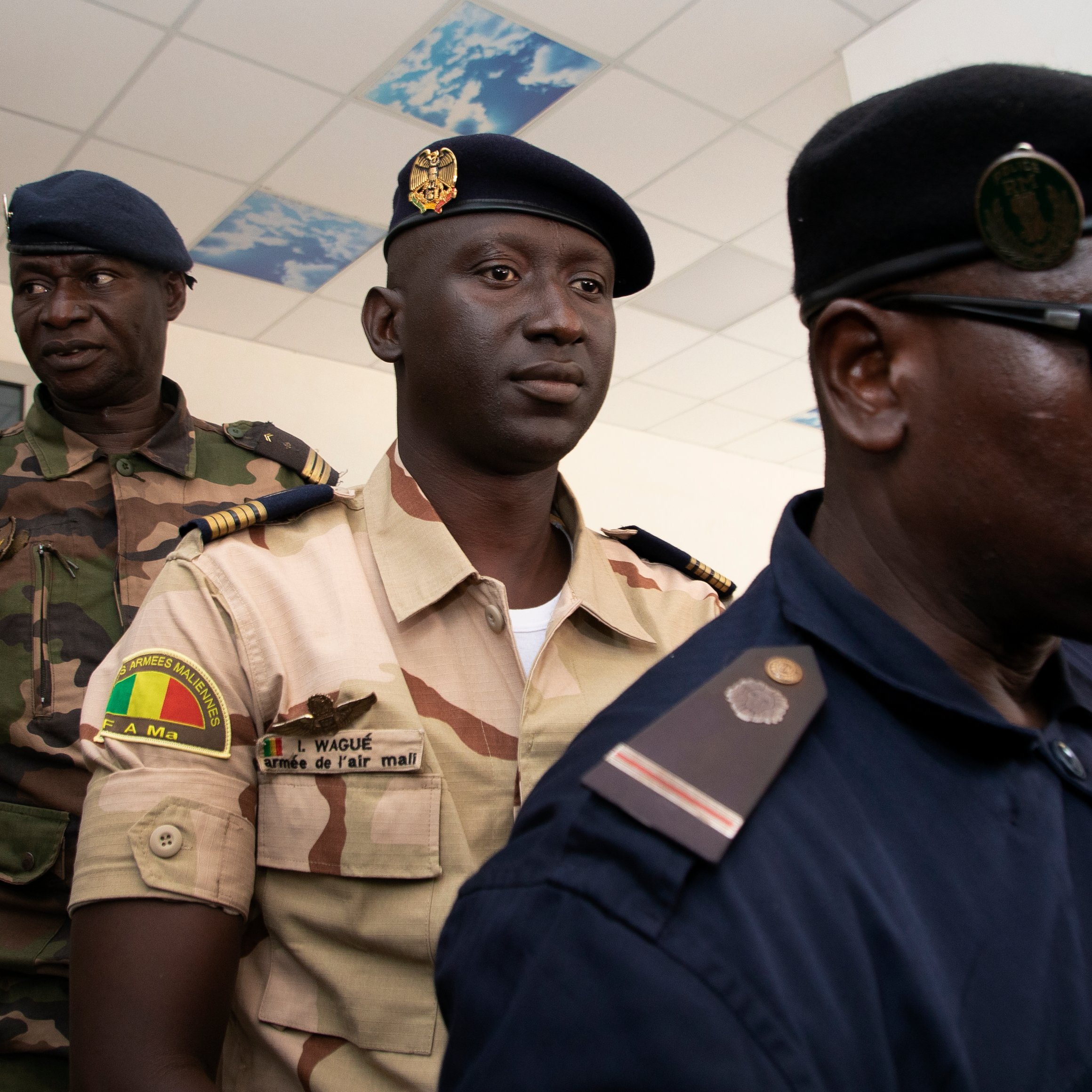 L'image montre un groupe d'hommes en uniforme, probablement des membres des forces de sécurité. Au premier plan, on peut voir un homme en uniforme sombre, portant un chapeau, qui parle à un microphone. Il semble être en train de faire une déclaration ou une interview. À l'arrière-plan, deux autres hommes en uniforme sont présents. L'un d'eux porte un uniforme plus clair avec des insignes, tandis que l'autre a un uniforme de camouflage. L'environnement semble être un bâtiment officiel, avec des murs clairs et un plafond décoré. L'atmosphère peut être perçue comme sérieuse et formelle.