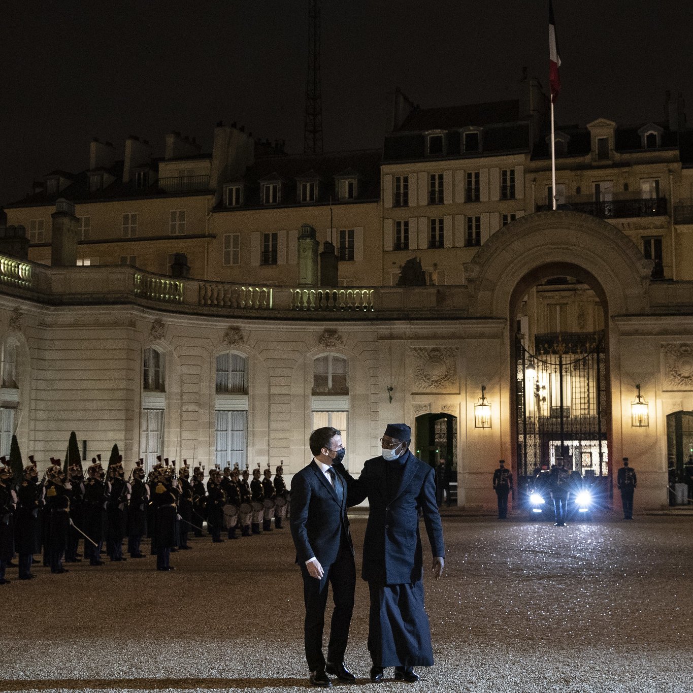 L'image représente une scène nocturne au Palais de l'Élysée, en France. Au premier plan, deux hommes discutent, l'un portant un costume noir et l'autre vêtu d'un long manteau sombre. En arrière-plan, une rangée de soldats en uniforme se tient en position, formant une haie d'honneur. Les bâtiments classiquement architecturés du palais sont illuminés, mettant en valeur leurs détails. Une lumière provenant d'un véhicule est visible à droite, ajoutant à l'atmosphère solennelle du moment. Les drapeaux français flottent au-dessus du bâtiment. L'ensemble de la scène dégage une impression de formalité et de dignité.