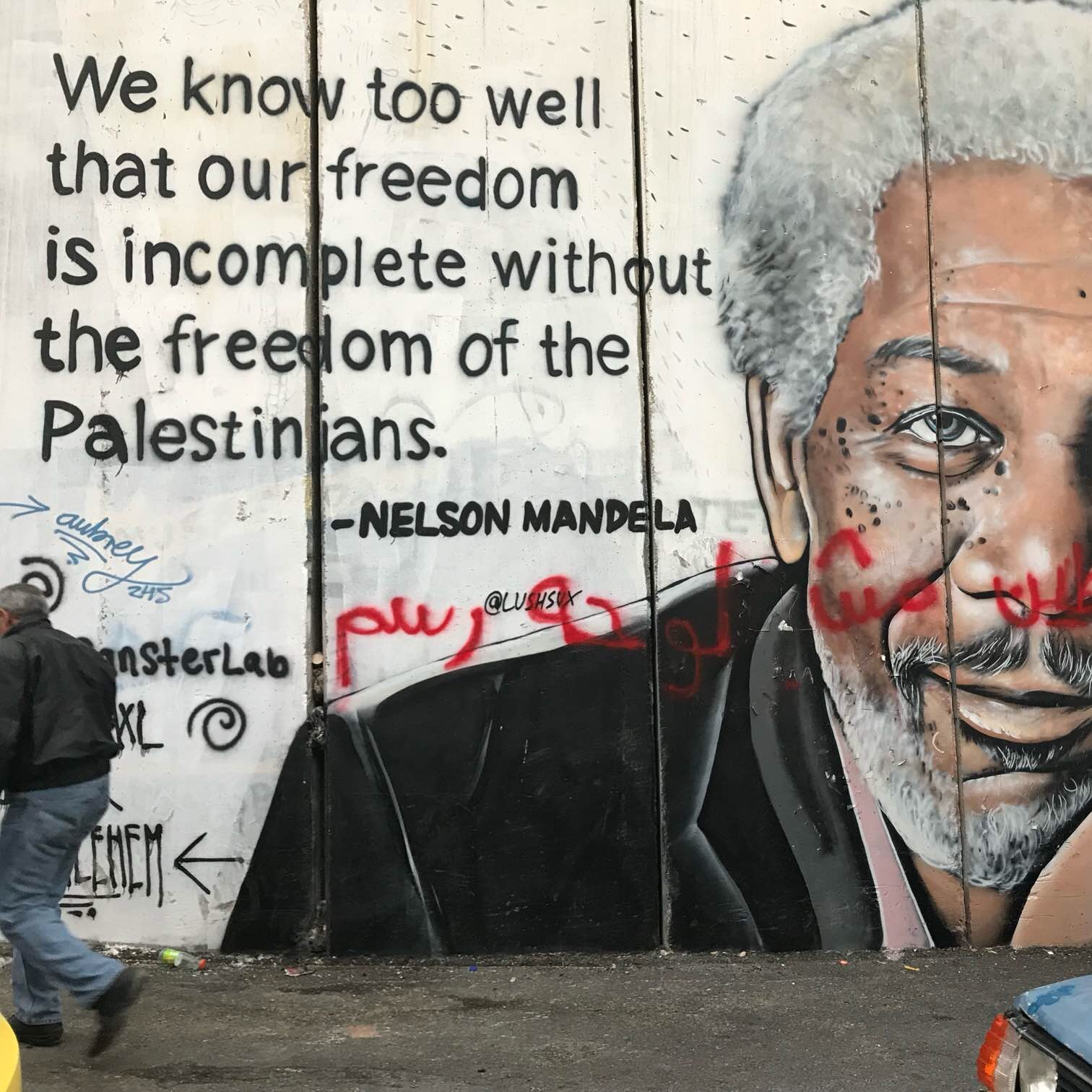 The image depicts a mural on a concrete wall, featuring a portrait of a man with gray hair and a thoughtful expression. The mural also includes a quote attributed to Nelson Mandela that reads, "We know too well that our freedom is incomplete without the freedom of the Palestinians." The background is a textured concrete surface, and there are some graffiti elements in red. The scene suggests themes of solidarity and freedom, particularly regarding the Palestinian struggle.