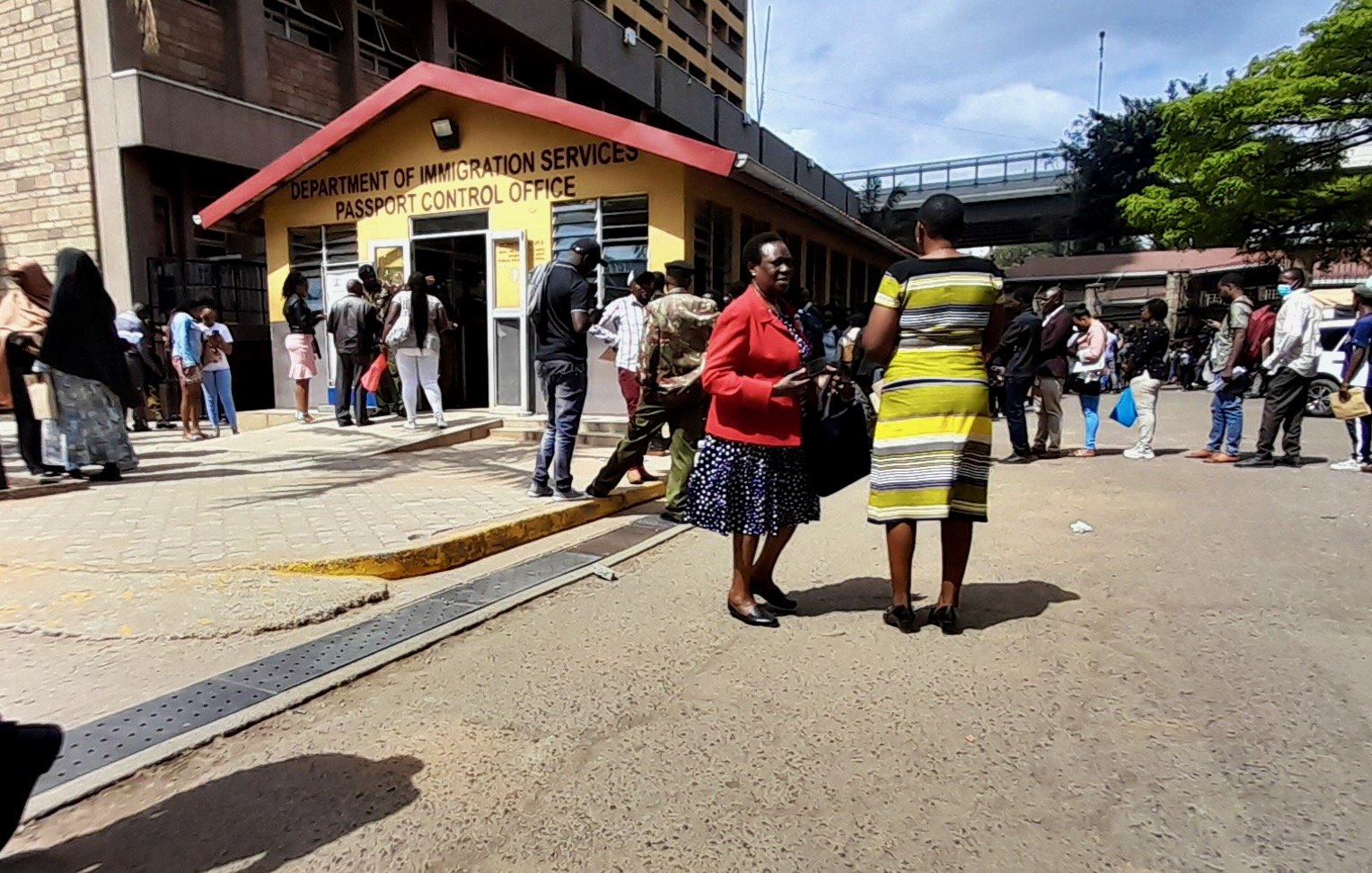 L'image montre une scène animée à l'extérieur d'un bureau de contrôle des passeports, identifié comme le "Department of Immigration Services". À l'avant-plan, deux femmes discutent, l'une portant une jupe et un haut noir avec des motifs, et l'autre vêtue d'une robe rayée jaune. L'environnement est urbain, avec des bâtiments en arrière-plan et une foule de personnes qui attendent ou se déplacent, créant une ambiance dynamique. Le ciel est partiellement nuageux, et l'atmosphère semble être celle d'une journée active. Les surfaces sont en béton, ajoutant un aspect concret à l'ensemble de la scène.