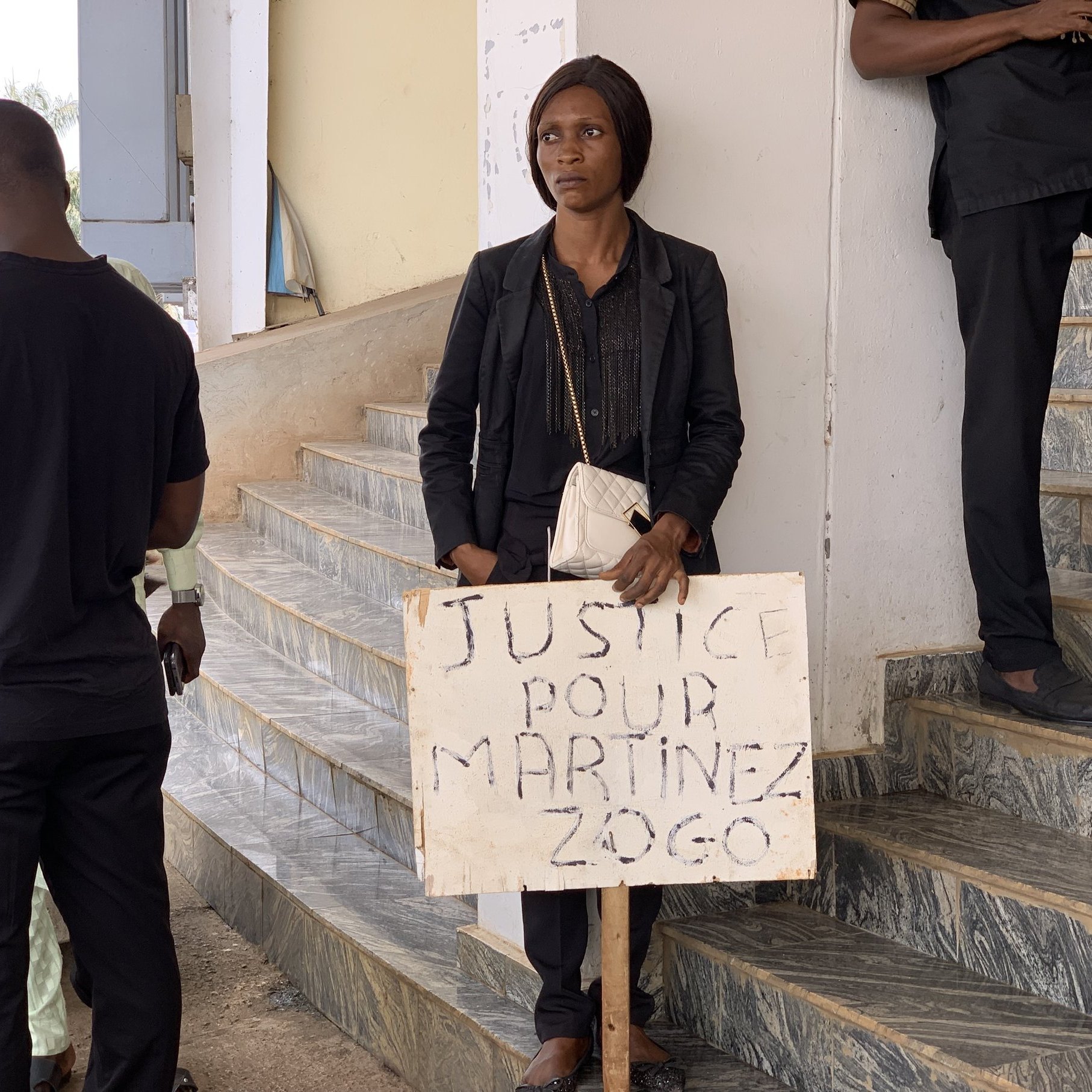 Dans cette image, on voit une femme debout dans un espace public. Elle porte des vêtements noirs et tient un panneau en bois qui porte l'inscription "JUSTICE POUR MARTINEZ ZOGO". La femme a une expression sérieuse et semble déterminée. À proximité, plusieurs personnes en vêtements noirs se tiennent, suggérant un rassemblement ou une manifestation. L'environnement est urbain, avec des escaliers visibles et des bâtiments en arrière-plan, indiquant un cadre où des actions sociales ou des revendications ont lieu.