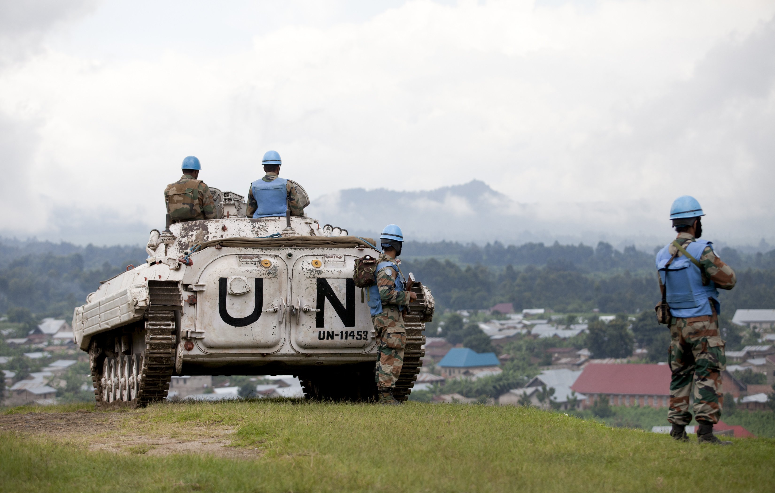 L'image présente un paysage où des soldats en uniforme de l'ONU se trouvent autour d'un véhicule blindé. Le véhicule, marqué par le symbole "UN" en lettres blanches, est stationné sur une colline qui surplombe une petite ville. Les soldats portent des casques bleu clair typiques des missions de maintien de la paix. À l'arrière-plan, on aperçoit des maisons aux toits colorés et une végétation dense, tandis que le ciel est partiellement nuageux, créant une atmosphère sereine malgré le contexte militaire. Les soldats semblent vigilants et concentrés, surveillant les environs.