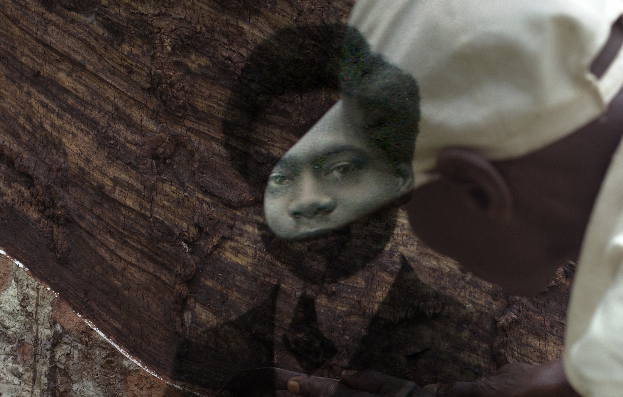 L'image montre un homme en train de travailler avec un morceau de bois. Ce dernier est une surface très texturée, et l'homme semble utiliser un outil pour façonner ou sculpter le bois. À travers le bois, un portrait en noir et blanc de quelqu'un d'autre est visible, créant un effet de superposition. Le visage qui apparaît dans le bois a une expression sérieuse, et l'homme qui travaille porte une casquette. L'atmosphère de l'image évoque une connexion entre le passé et le présent, comme si l'artisan essayait de donner vie à la mémoire de cette personne.