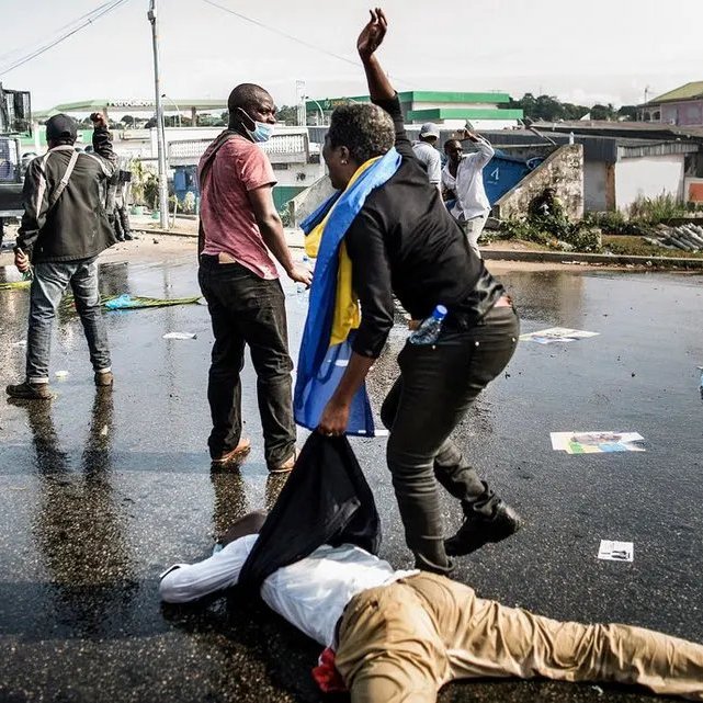 L'image montre une scène de manifestation dans une rue. Au premier plan, on peut voir des personnes en mouvement, certaines en train de gesticuler, tandis qu'une personne est à terre, apparemment blessée. L'un des manifestants porte un drapeau, probablement symbolique. En arrière-plan, il y a un véhicule de police, ce qui suggère une présence des forces de l'ordre. Le sol est couvert de débris, comme des affiches et des bouteilles, témoignant de la tension de la situation. L'ambiance semble intense et sérieuse, avec une impression de conflit.