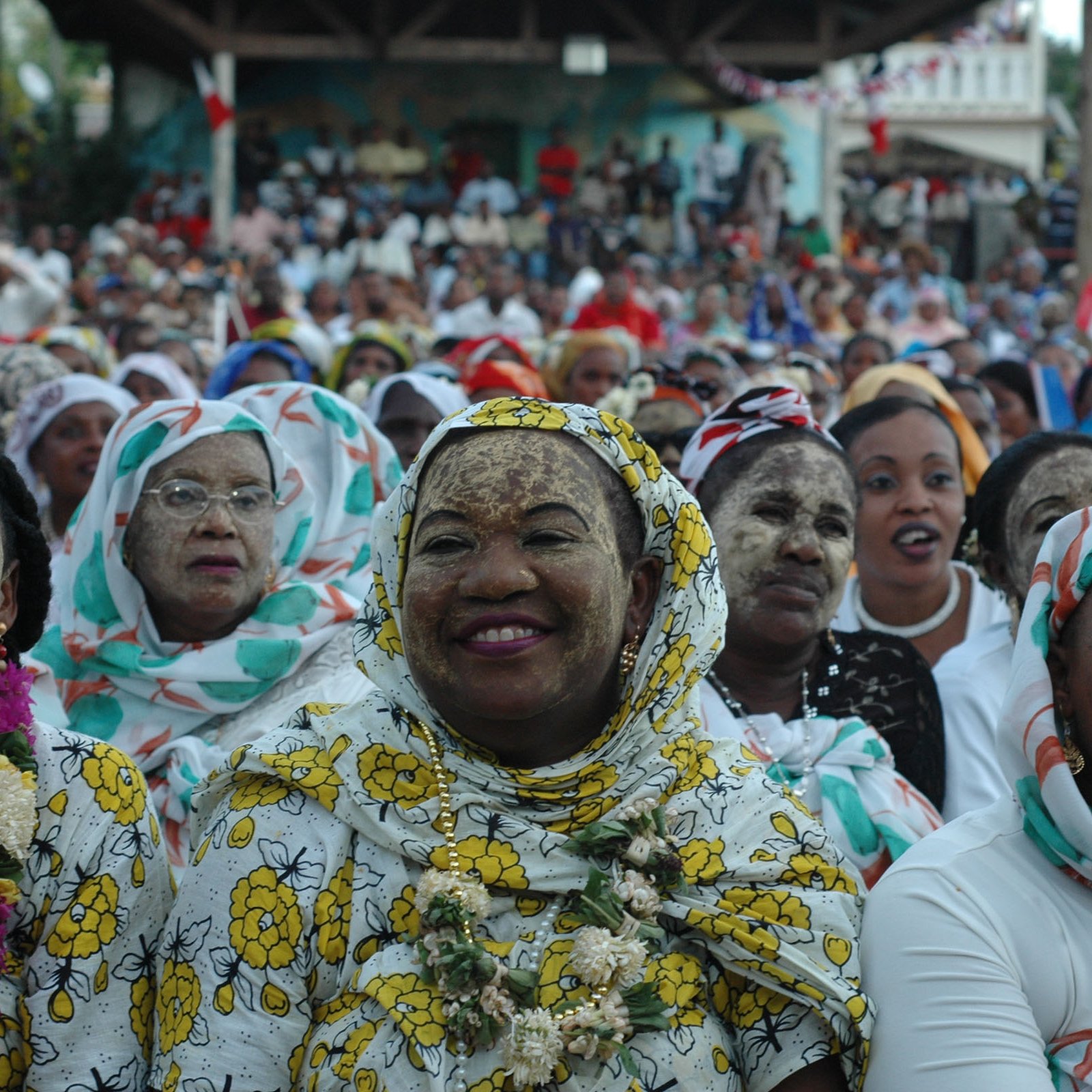 L'image montre un large groupe de personnes réunies pour un événement, probablement festif. Au premier plan, des femmes portent des vêtements colorés et traditionnels, agrémentés de motifs floraux. Certaines ont le visage recouvert de farine ou d'une substance claire, créant une texture particulière. Les femmes semblent joyeuses, souriant et engaged dans l'événement. En arrière-plan, l'assistance s'étend jusqu'à une grande scène, suggérant une atmosphère festive vibrante. Les couleurs et les motifs des vêtements ajoutent une dimension culturelle à l'ensemble de la scène. L'ambiance générale est celle d'une célébration communautaire, remplie de vie et de convivialité.