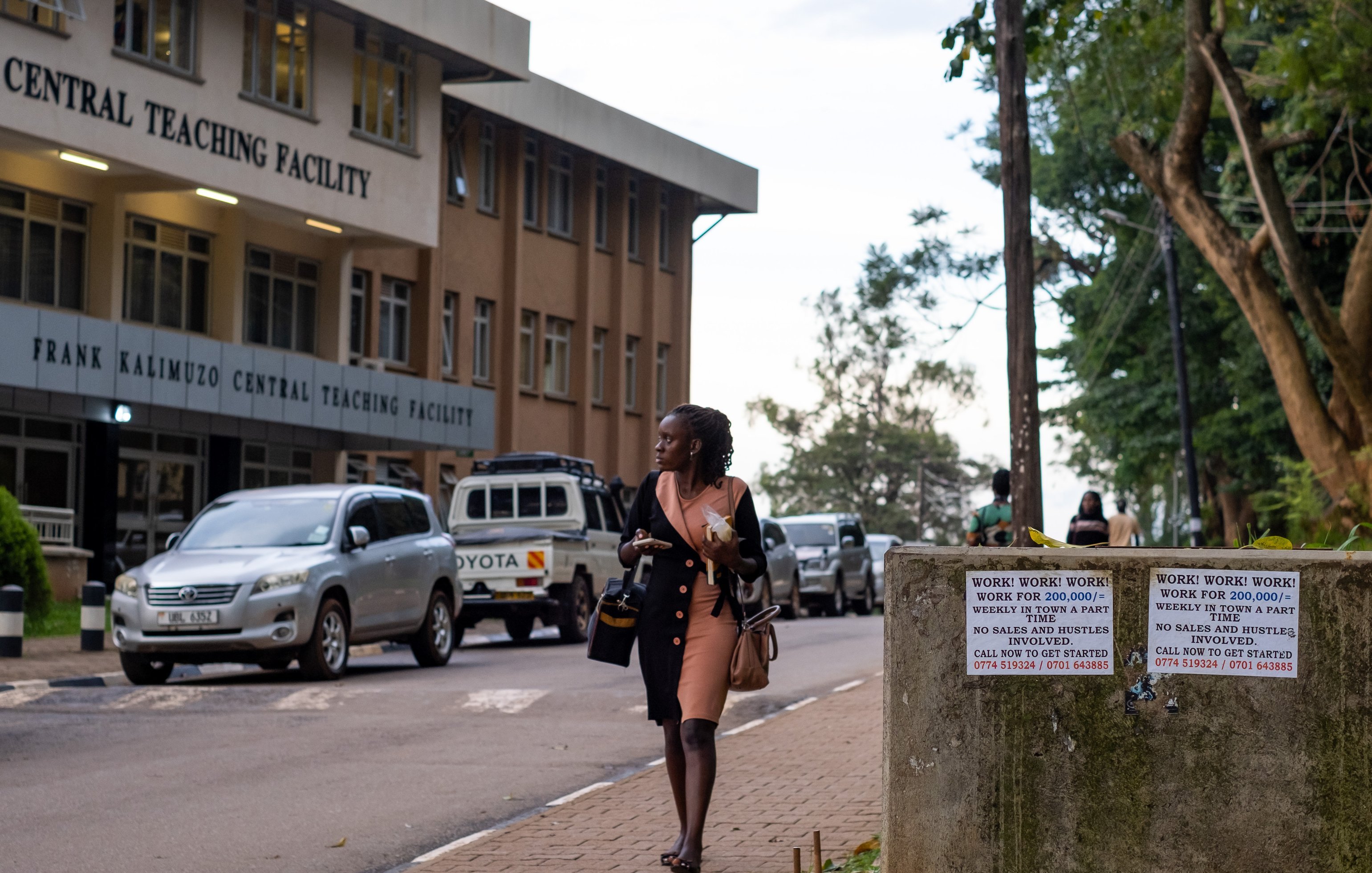 Dans cette image, on voit une scène urbaine près d'un bâtiment nommé "Frank Kalimuzo Central Teaching Facility". La structure est moderne avec plusieurs fenêtres et un design clair. Sur le trottoir, une femme vêtue d'une robe rose marche, portant une sacoche. À l'arrière-plan, on aperçoit quelques véhicules, dont une voiture blanche, et des arbres qui apportent une touche de verdure à l'environnement. Des panneaux sur un mur en béton à droite attirent l'attention avec des avertissements écrits en grandes lettres. L'ambiance est calme, suggérant un moment de transition entre la journée et le soir.