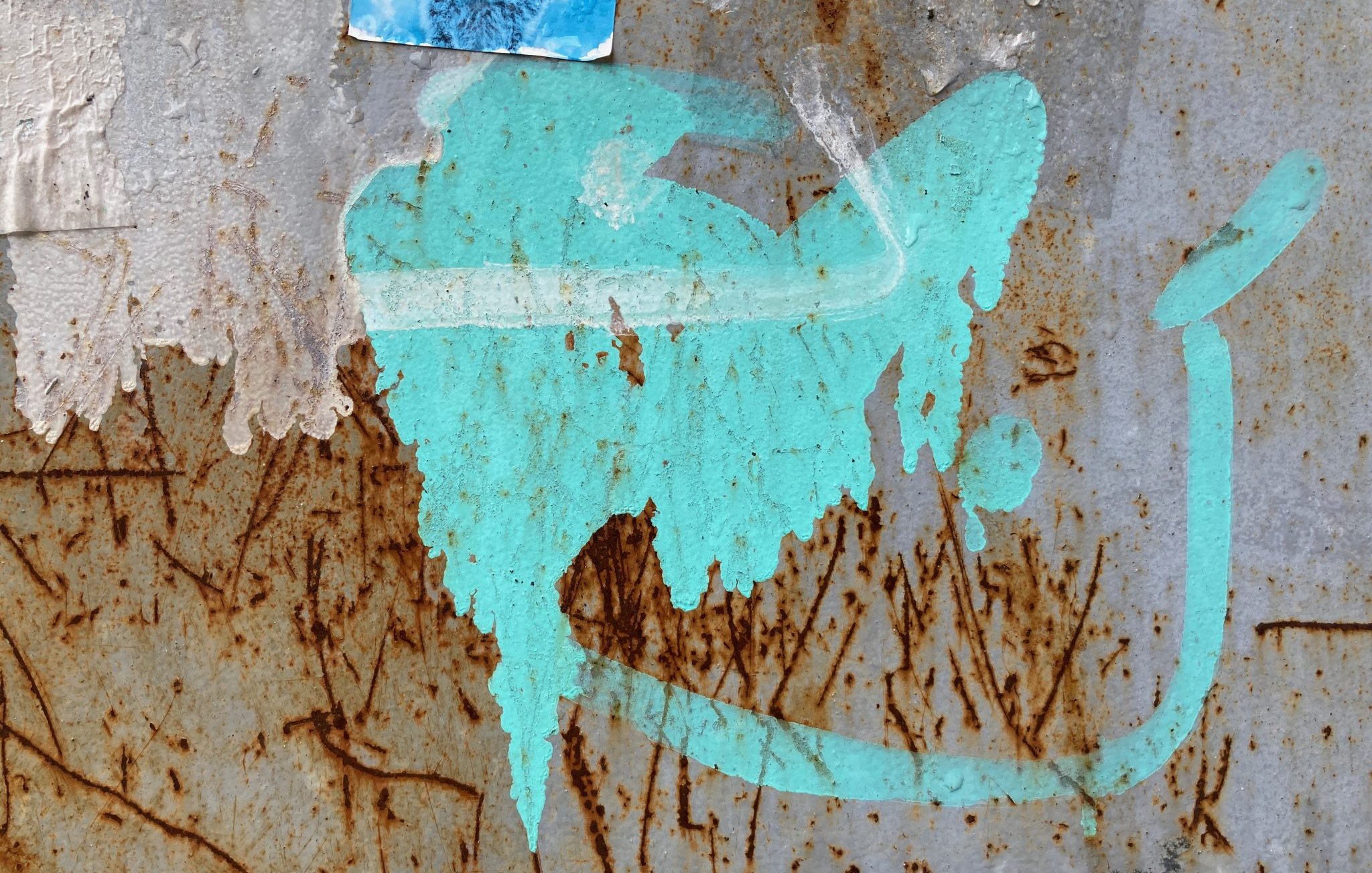 L'image présente une surface en métal, probablement rouillée, avec des marques et des textures variées. On y voit une tache de couleur turquoise qui semble avoir été projetée. Cette tache a une forme fluide et irrégulière, évoquant une sorte de mouvement ou de dynamisme. À proximité, on peut distinguer des traces brunes, résultant de la rouille, qui ajoutent un contraste marqué à la couleur vive. Le fond est texturé, avec des stries et des imperfections, suggérant l'âge et l'usure du matériau. L'ensemble crée une atmosphère à la fois urbaine et artistique.