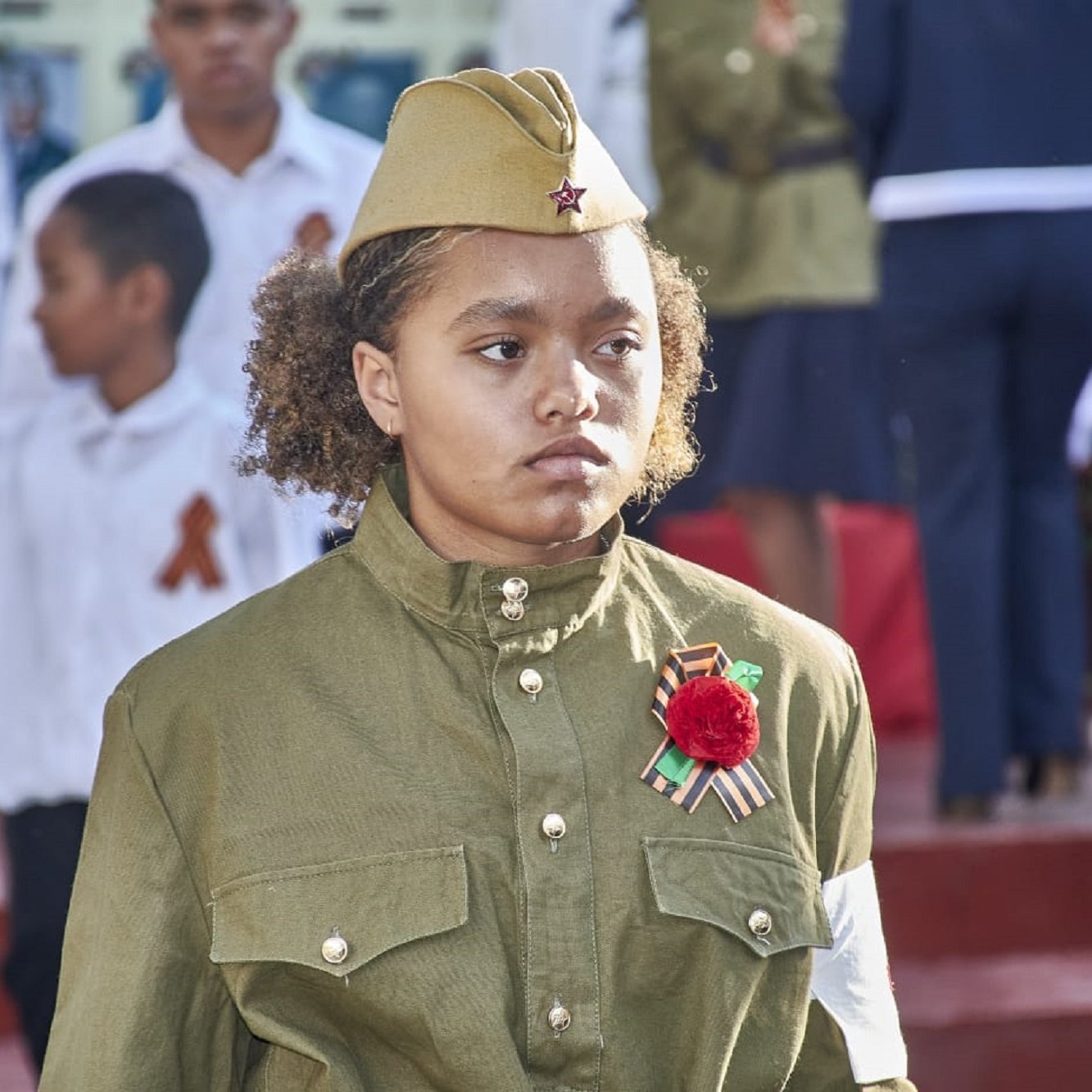 L'image montre une jeune fille portant un uniforme militaire, qui est de couleur olive et a des ornements spécifiques. Elle porte un chapeau similaire, typique des uniformes militaires. Sur sa poitrine, elle affiche une décoration, probablement une médaille ou un insigne, ainsi qu'une fleur rouge. En arrière-plan, on aperçoit d'autres personnes, dont des enfants en vêtements blancs et d'autres habits festifs, suggérant une célébration ou un événement commémoratif. L'expression de la jeune fille semble sérieuse, soulignant l'importance du moment. Le cadre est marqué par une ambiance solennelle, car plusieurs personnes semblent participer à une cérémonie.