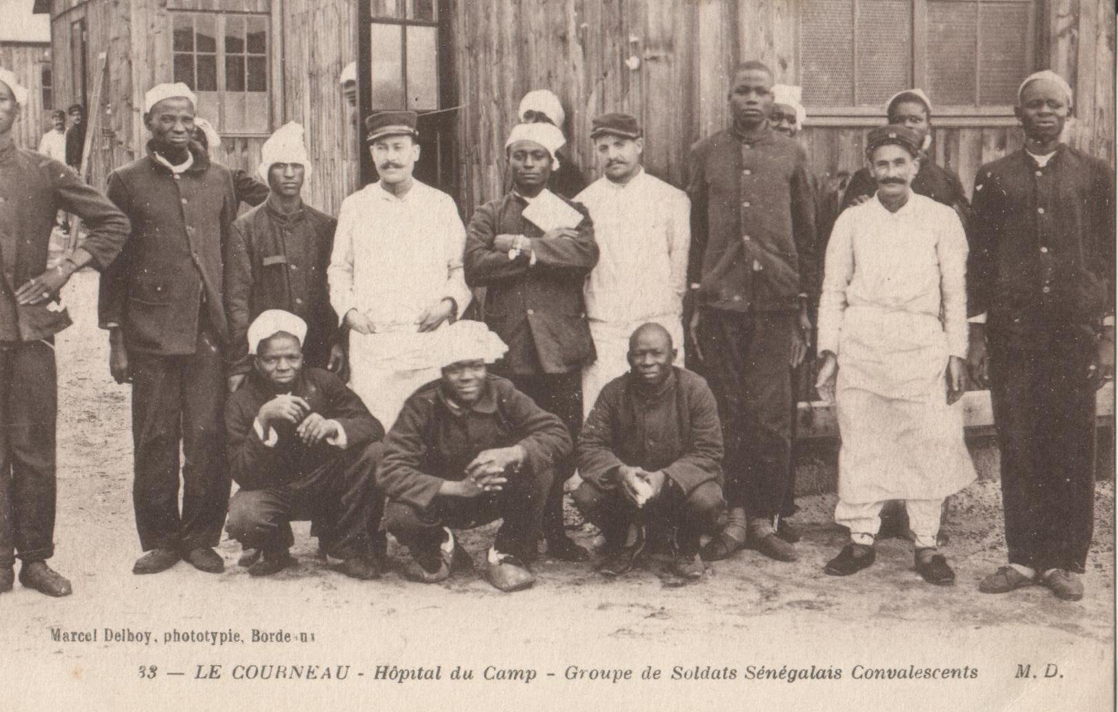 L'image montre un groupe de soldats sénégalais convalescents rassemblés devant un bâtiment en bois, vraisemblablement un hôpital de campagne. Il y a treize personnes, dont des hommes en uniforme militaire et des membres du personnel médical. Les soldats portent des uniformes sombres et certains d'entre eux ont des casquettes ou des bonnets. Le personnel médical, d'autre part, porte des blouses blanches, indiquant leur rôle. Tous affichent des expressions amicales, certains sourient tandis que d'autres prennent des poses plus sérieuses. L'arrière-plan montre des murs en bois, donnant une atmosphère de campagne ou de mobilisation militaire. Cette scène capture un moment de camaraderie et de résilience au sein des soldats pendant une période difficile.