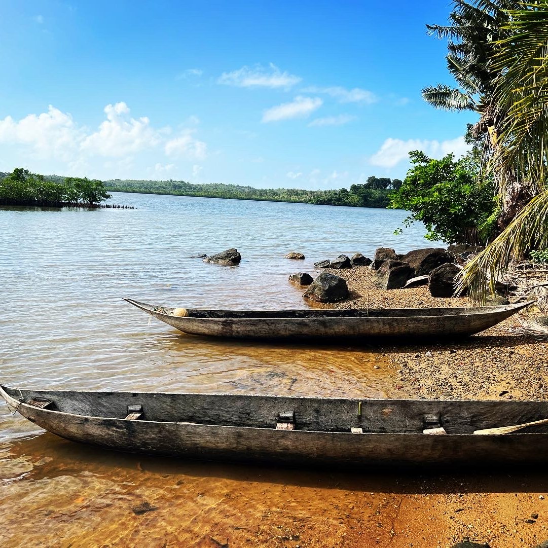 Imaginez une scène paisible au bord de l'eau. Devant vous se trouve un rivage doux avec du sable doré, illuminé par le soleil. À votre gauche, deux pirogues en bois, usées par le temps, reposent tranquillement sur le rivage, légèrement inclinées vers l'eau calme. La surface de l'eau est lisse, reflétant le ciel bleu parsemé de quelques nuages blancs. À droite, vous pouvez percevoir des rochers de tailles variées, surmontés ici et là de végétation luxuriante. Des palmiers se dressent en arrière-plan, ajoutant une note tropicale à l'ambiance. L'air est frais, avec une légère brise qui apporte une odeur de terre humide et de végétation. L'ensemble dégage une atmosphère de sérénité et de beauté naturelle, propice à la contemplation.