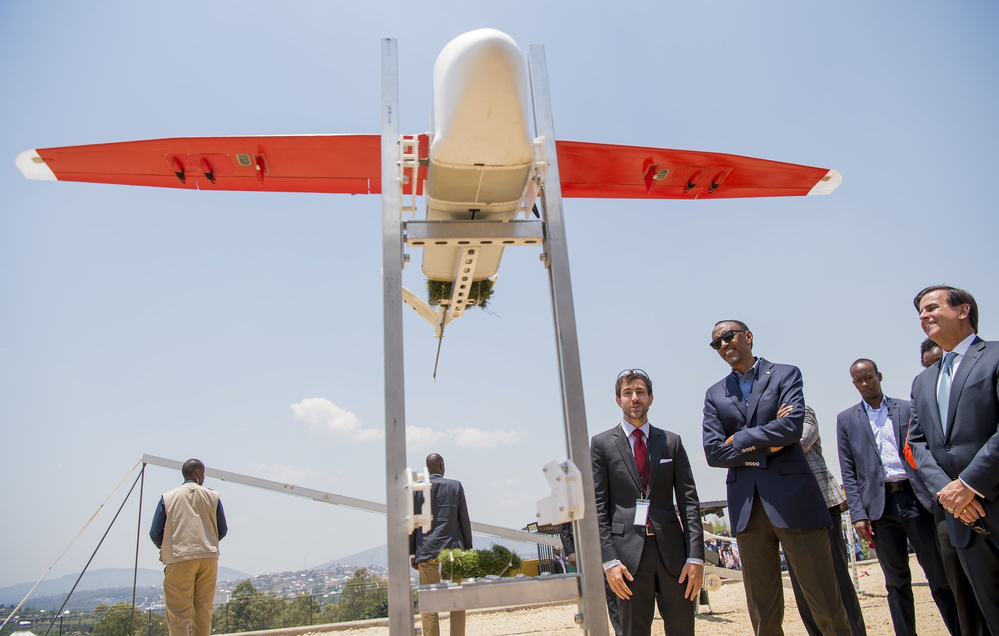 L'image montre un drone de forme allongée, aux ailes rouges et au corps blanc, placé sur une structure métallique. Plusieurs personnes, notamment des hommes en costumes, se tiennent à proximité, observant le drone avec intérêt. En arrière-plan, on aperçoit un paysage urbain et des collines sous un ciel bleu lumineux. L'atmosphère semble à la fois sérieuse et enthousiaste, suggérant un événement lié à la technologie ou à l'innovation.