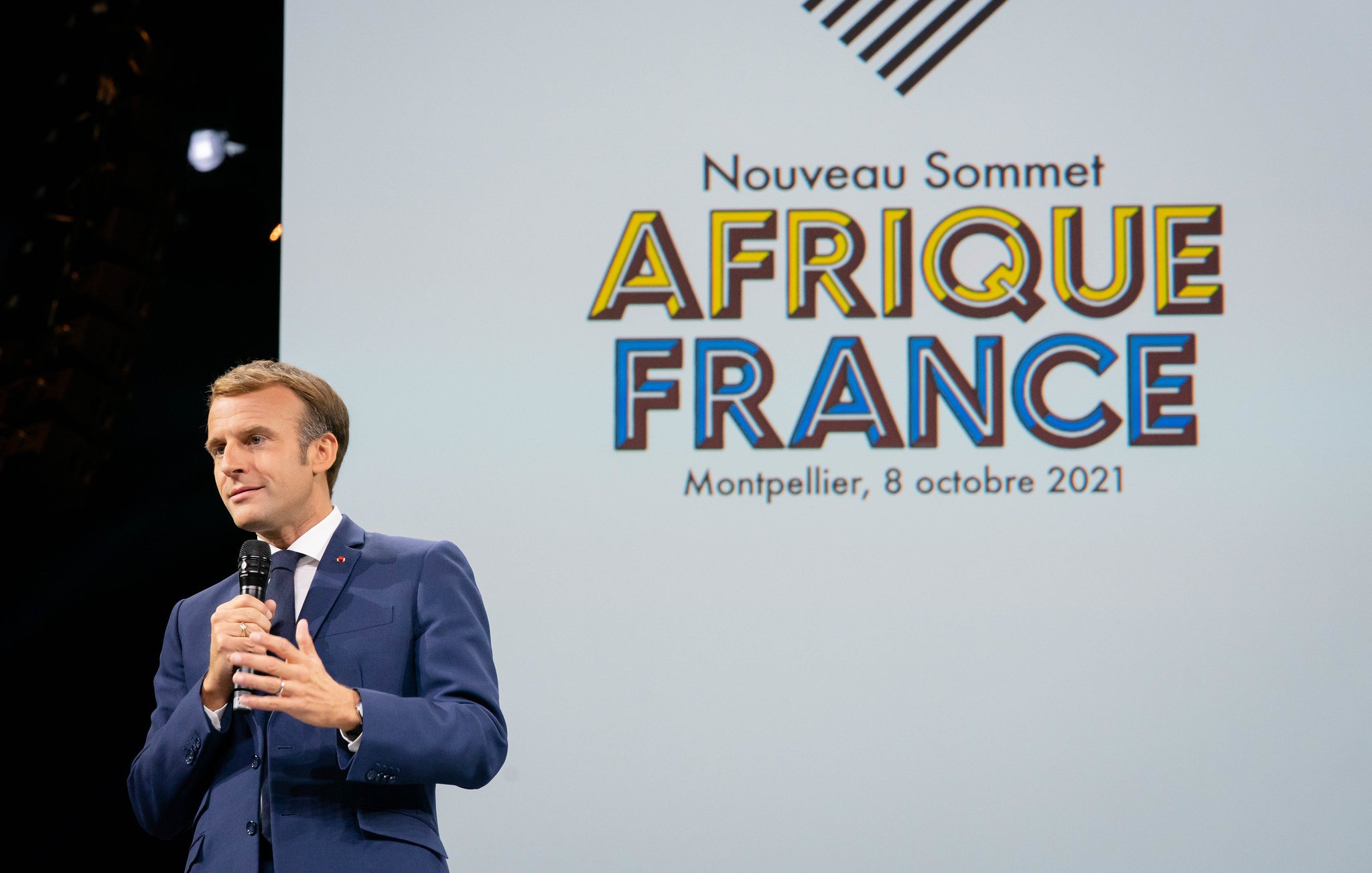 L'image montre un homme, probablement un leader ou un politicien, en train de s'adresser à un public. Il porte un costume sombre et une cravate, et il tient un micro dans sa main. En arrière-plan, il y a un grand écran avec le texte "Nouveau Sommet AFRIQUE FRANCE" suivi de la mention "Montpellier, 8 octobre 2021". L'ambiance est formelle et l'accent est mis sur la rencontre entre la France et les pays africains. Le public semble attentif à son discours.