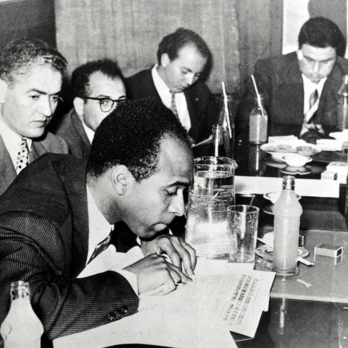 L'image en noir et blanc montre une scène de réunion dans un environnement intérieur, probablement une salle de conférence. Plusieurs hommes sont assis autour d'une table. Au premier plan, un homme est penché sur une feuille de papier, prenant semble-t-il des notes. Il est vêtu d'un costume sombre et d'une cravate, et semble concentré. À sa droite, un autre homme regarde vers le bas, tandis que les autres hommes en arrière-plan discutent ou examinent des documents. Sur la table, on peut voir des verres, des bouteilles, et quelques objets éparpillés. L'atmosphère semble sérieuse et studieuse, évoquant un moment important de collaboration ou de décision.