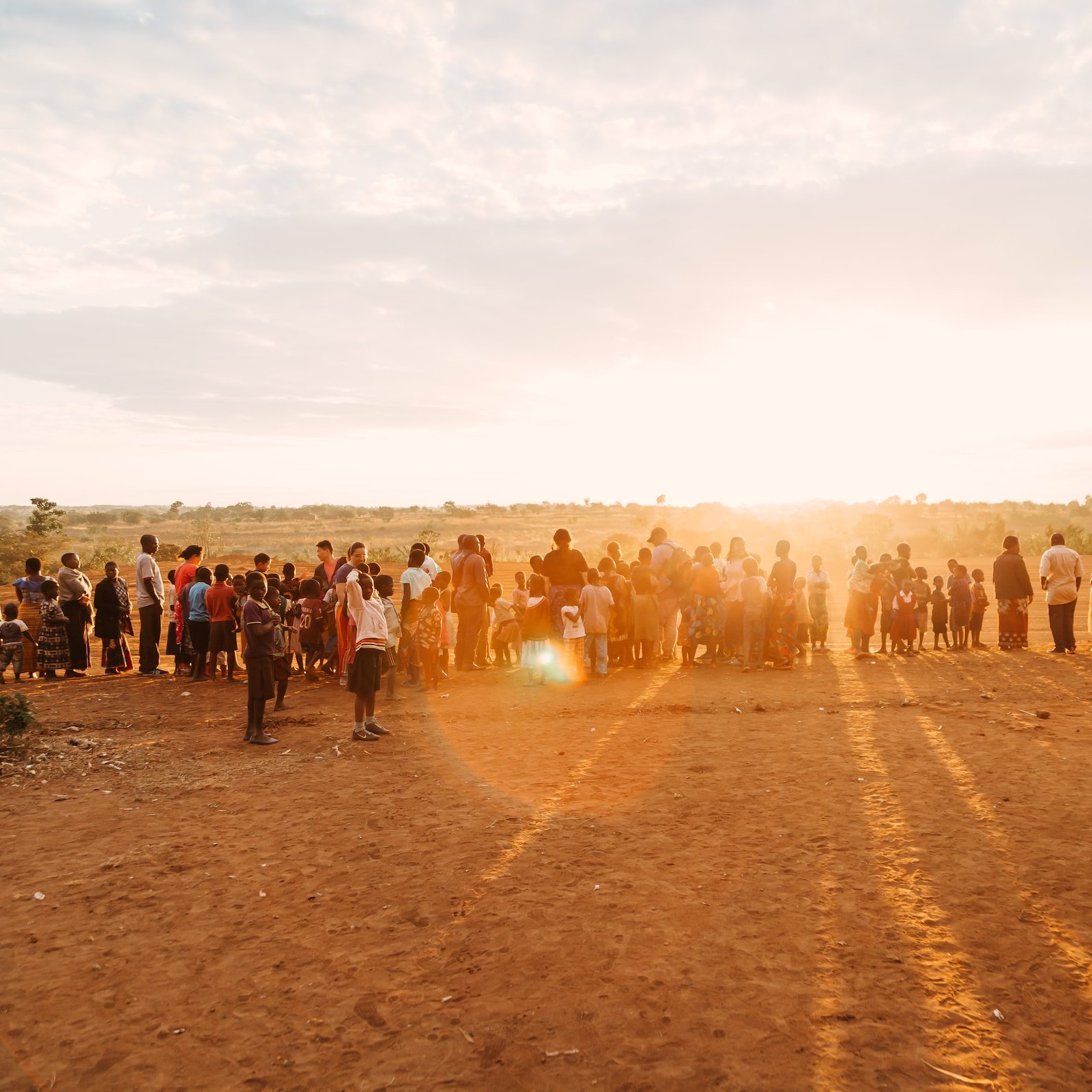 L'image présente une scène de rassemblement en plein air, au coucher du soleil. Au premier plan, un groupe nombreux de personnes se tient sur un terrain poussiéreux, probablement un village ou une communauté rurale. Les silhouettes des adultes et des enfants se distinguent distinctement alors qu'ils font face à l'horizon où le soleil descend, projetant de longues ombres sur le sol. L'arrière-plan est composé de plantes et d'une vaste étendue de paysage, créant une atmosphère paisible et chaleureuse. Le ciel, teinté de couleurs douces, suggère une fin de journée tranquille.