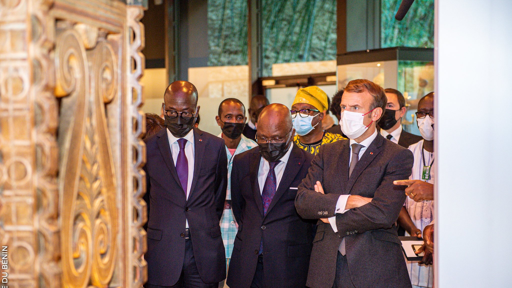 L'image montre un groupe de personnes, dont trois hommes, en train d'observer un objet d'art ou une exposition. Les hommes sont habillés de costumes formels, avec deux en costumes sombres et un en costume gris avec une cravate. Ils affichent des expressions de concentration et d'intérêt. À l'arrière-plan, on perçoit des éléments culturels, probablement des œuvres d'art ou des artifacts, créant une ambiance muséale. Les visiteurs portent tous des masques, ce qui indique qu'ils se trouvent dans un contexte où cela est nécessaire, peut-être en raison de préoccupations sanitaires. Le cadre est bien éclairé, mettant en valeur l'importance de l'exposition.