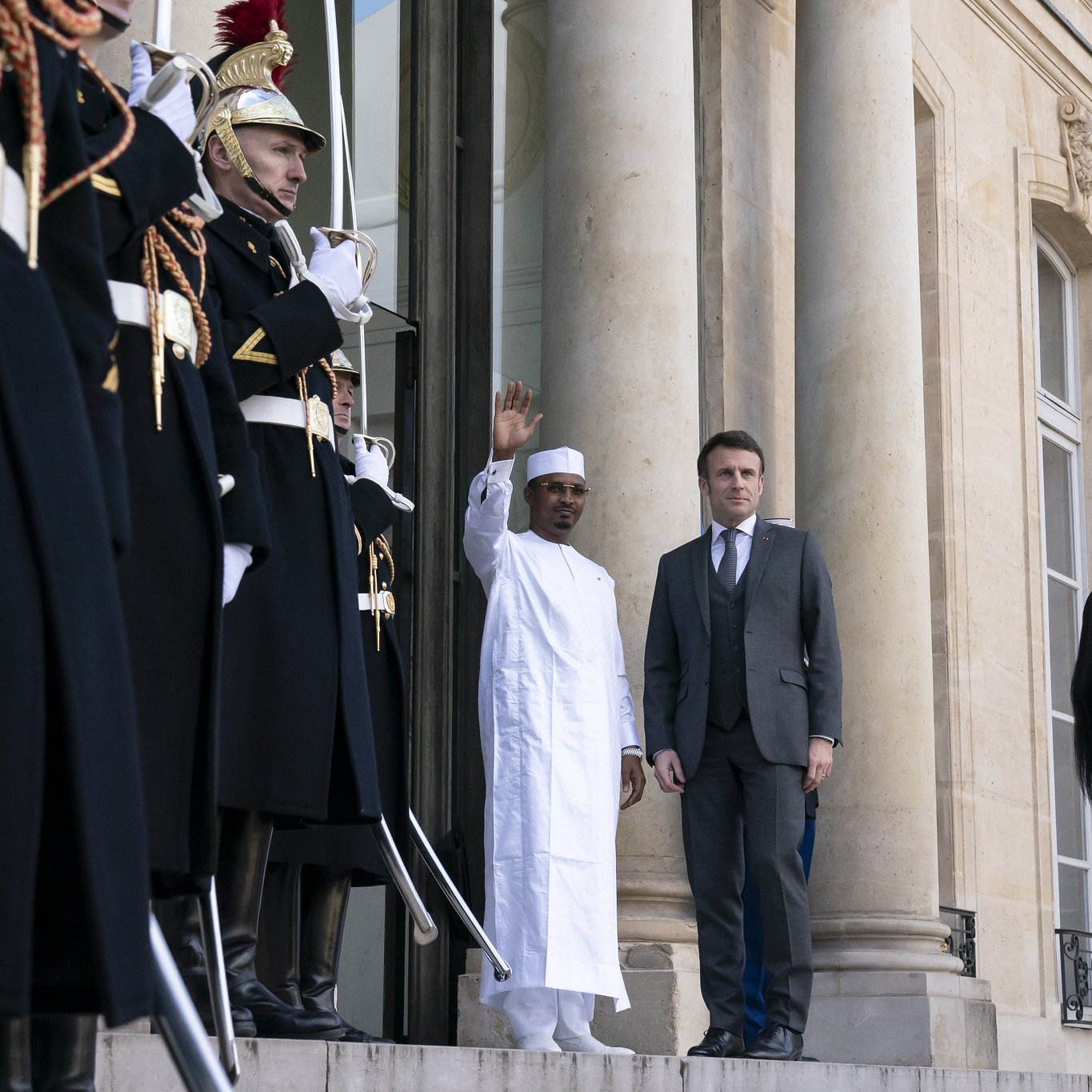 L'image montre une scène officielle à l'extérieur d'un bâtiment majestueux. Deux hommes se tiennent sur les marches devant l'entrée principale. À gauche, un homme porte une tenue traditionnelle blanche, levant la main en salutation. À sa droite, un homme en costume sombre regarde légèrement vers lui. En arrière-plan, plusieurs gardes d'honneur, habillés de costumes noirs avec des ornements dorés et des plumes rouges sur leurs casques, se tiennent en position, ajoutant à l'atmosphère solennelle de la scène. Les colonnes imposantes du bâtiment et les détails architecturaux mettent en valeur le caractère formel de l'événement.