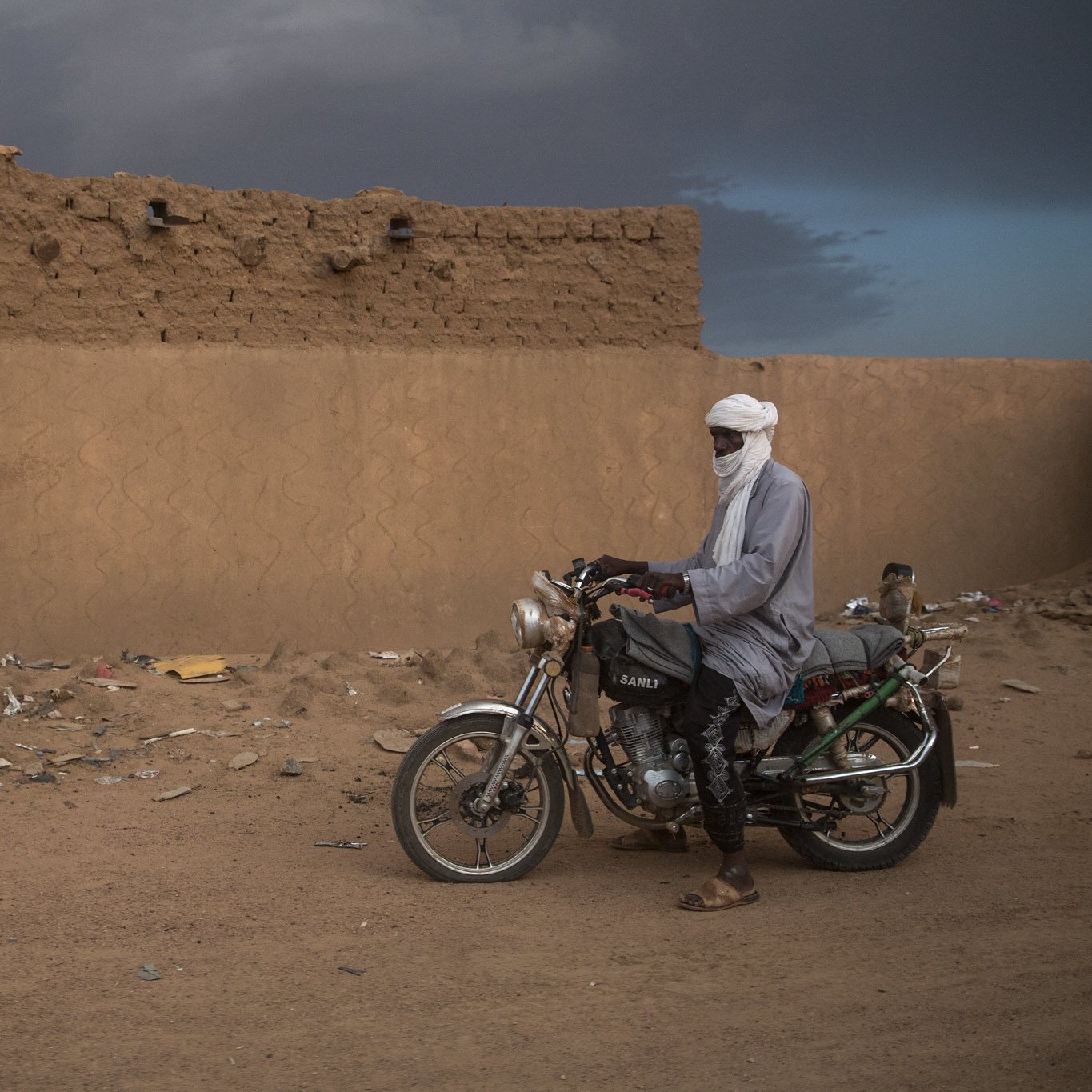 L'image montre un homme immobile à côté de sa moto dans un paysage désertique. Il porte un long vêtement traditionnel et un turban qui couvre sa tête, laissant seulement ses yeux visibles. Le sol est constitué de sable et de débris, tandis qu'en arrière-plan, on distingue un mur de terre crue, simple et robuste, typique des constructions dans cette région. Le ciel est nuageux, ajoutant une atmosphère dramatique à la scène. L'homme semble contemplatif, connectant ainsi l'élément humain avec l'environnement désertique qui l'entoure.