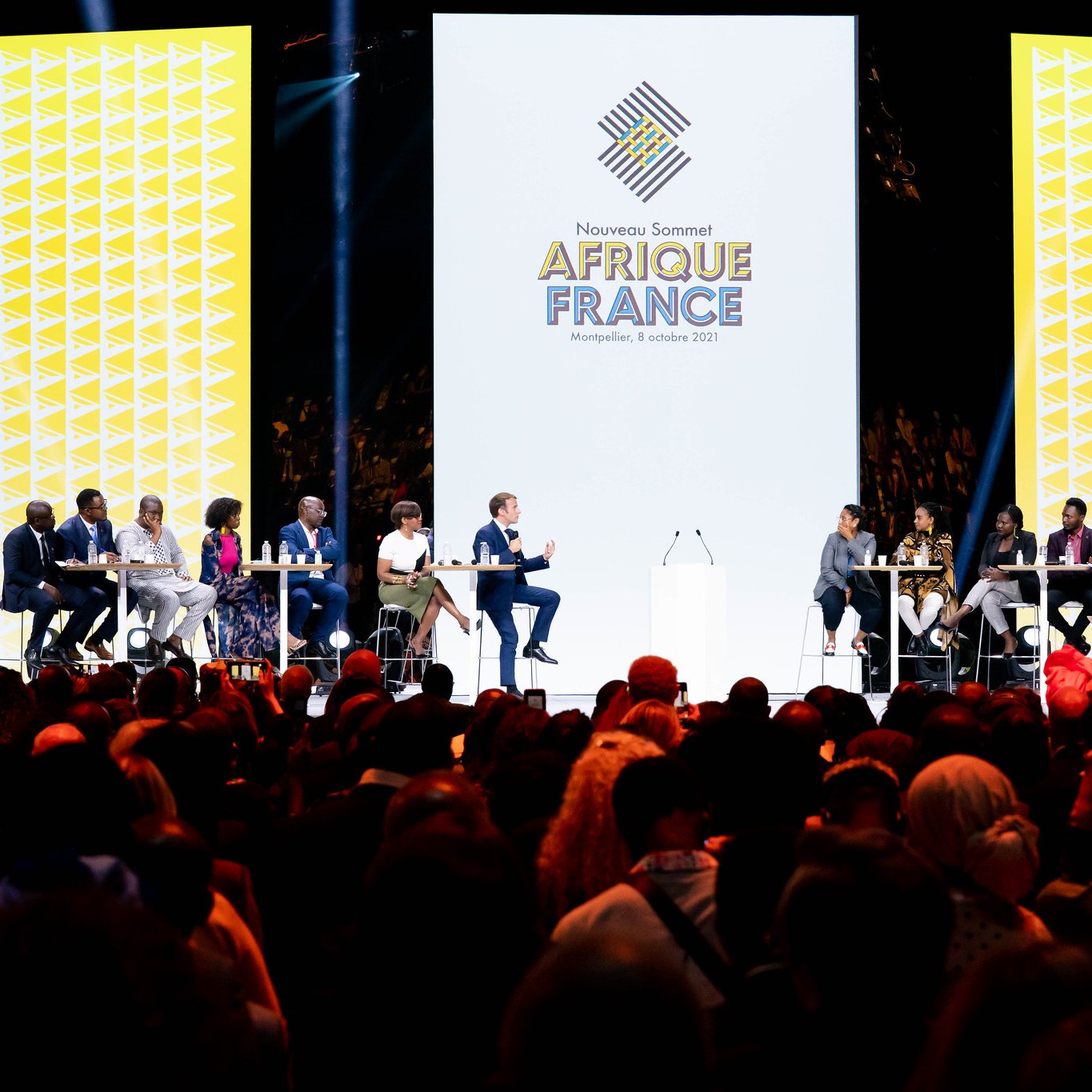 L'image montre une scène d'événement avec plusieurs intervenants assistant à un sommet intitulé "Afrique France". Au centre, un grand écran affiche le logo de l'événement avec une conception graphique moderne. Les intervenants, assis à des tables en cercle, semblent engagés dans une discussion. L'audience est nombreuse et variée, occupant une grande partie de l'espace devant la scène, créant une atmosphère dynamique et interactive. L'éclairage est focalisé sur la scène, mettant en valeur les participants.