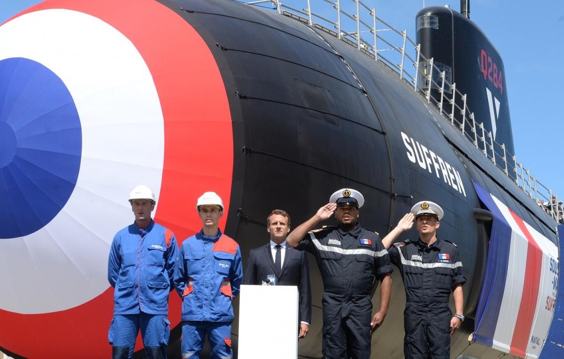 L'image montre un sous-marin de couleur noire avec un grand cercle rouge et bleu sur son côté. Devant le sous-marin, il y a quatre personnes en tenue de travail, dont deux en bleu et deux en uniforme noir. Elles se tiennent debout, affichant des expressions sérieuses. En arrière-plan, le ciel est clair et bleu, donnant une ambiance ensoleillée à la scène. Au centre, une plate-forme sur laquelle un homme en costume parle, probablement lors d'une cérémonie liée au sous-marin. Cela évoque une atmosphère de célébration ou d'inauguration d'un nouveau bateau.