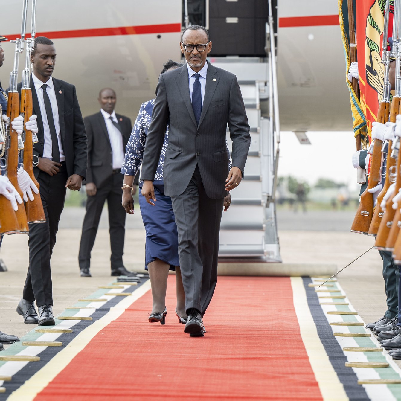 L'image montre un moment officiel sur un tapis rouge, où un homme en costume, probablement un dignitaire, marche avec assurance. Il est entouré de garde d'honneur en tenue formelle, tenant des fusils. À l'arrière-plan, on aperçoit une grande échelle d'avion et d'autres personnes en costume. L'atmosphère dégage un sentiment de cérémonie et de respect, soulignée par la disposition soignée des militaires et le faste du décor.