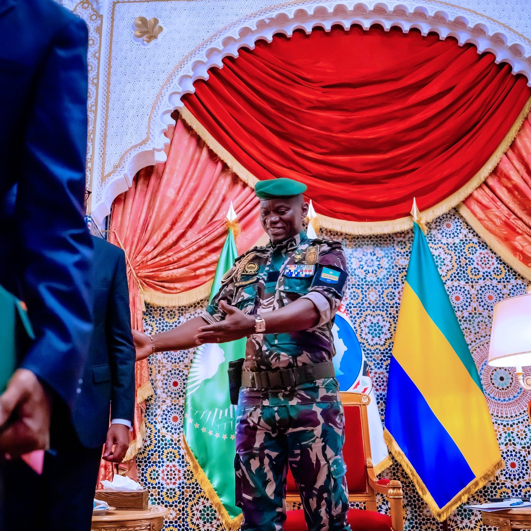 L'image montre un salon d'apparat coloré, avec un décor richement orné et des drapeaux en arrière-plan, dont on distingue le drapeau du Gabon. Au centre, un homme portant un uniforme militaire camouflé et une casquette verte se tient debout avec un sourire accueillant. Il est en train de tendre la main vers une autre personne, probablement pour une poignée de main. Les murs sont décorés de motifs arabes et la scène dégage une atmosphère de formalité et de respect. Une lampe est placée à droite, ajoutant une touche de chaleur à l'environnement.