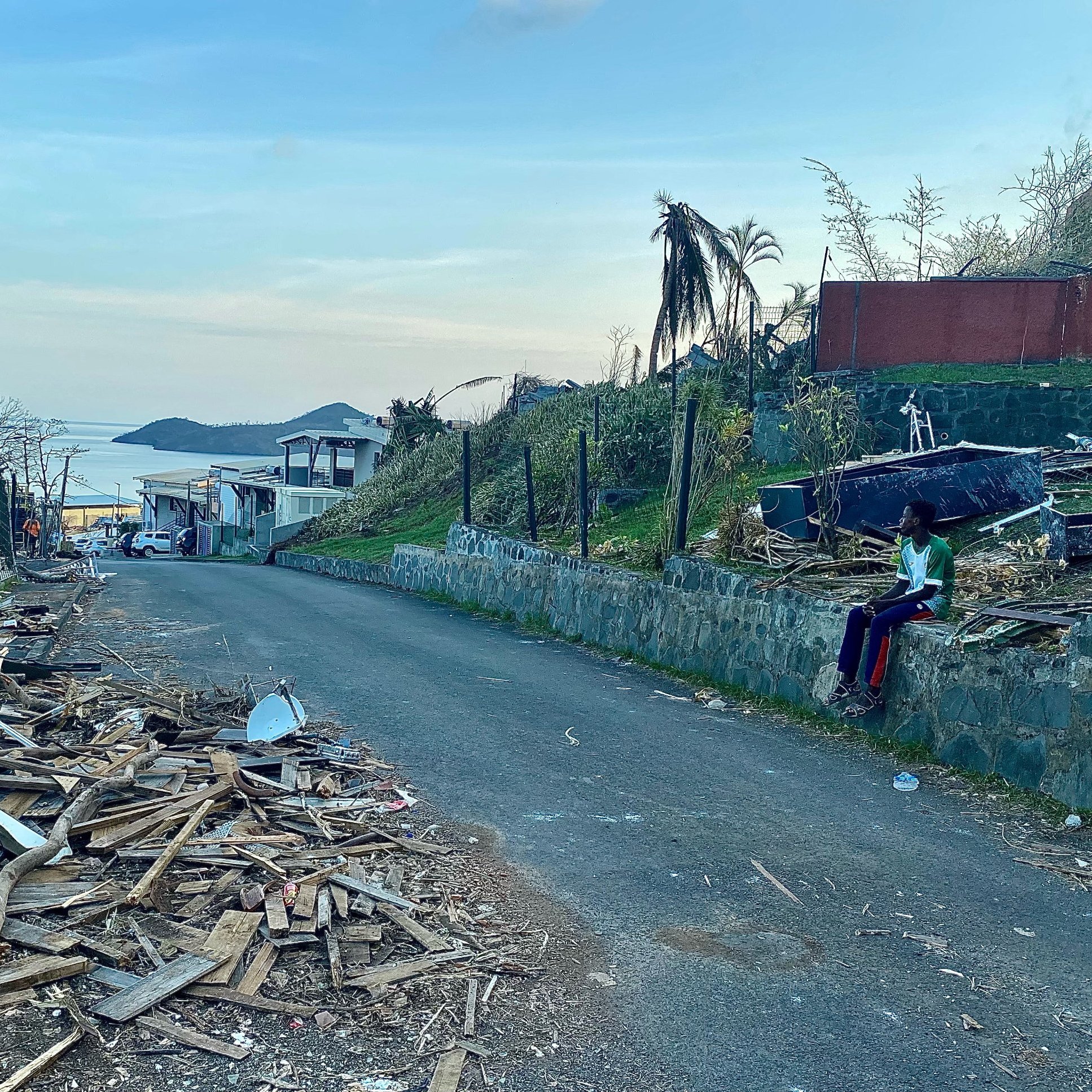 L'image présente une scène de destruction dans une rue. On voit des débris en bois éparpillés sur le sol, témoignant d'un événement dévastateur, peut-être une tempête ou un ouragan. Sur la gauche, il y a des maisons endommagées, certaines avec des toits manquants. En arrière-plan, on devine un paysage côtier avec une étendue d'eau qui reflète la lumière du ciel. À droite, une personne est assise sur un mur, habillée de vêtements colorés, semblant contempler les dégâts. La lumière du jour est douce, annonçant un crépuscule paisible, contrastant avec la morosité de la scène. Les palmiers, certains déracinés, ajoutent à l'impression de désolation.