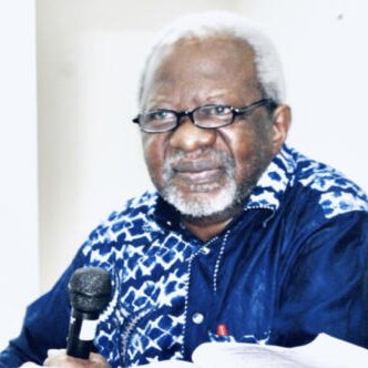 L'image présente un homme assis à une table, tenant un microphone. Il porte des lunettes et a des cheveux gris et bouclés. Son visage exprime une certaine sagesse et réflexion. Il est habillé d'une chemise à motifs, principalement bleue et blanche. Le fond de l'image est neutre, mettant en avant sa présence. On peut sentir une atmosphère de concentration, probablement lors d'un discours ou d'une discussion.