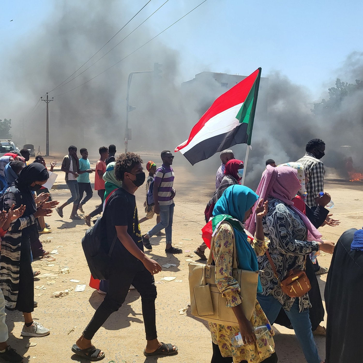 L'image montre une scène de manifestation animée. Des groupes de personnes marchent dans une rue, portant des drapeaux, dont un qui représente le Soudan. La foule paraît déterminée, et beaucoup de manifestants portent des masques, probablement en raison des conditions de pollution ou pour des raisons de sécurité. En arrière-plan, on peut voir des colonnes de fumée noire qui s'élèvent dans le ciel, créant une atmosphère tendue. Les manifestants semblent traverser une intersection, avec des véhicules garés sur le côté. L'atmosphère générale dégage une impression de forte mobilisation et de passion collective.
