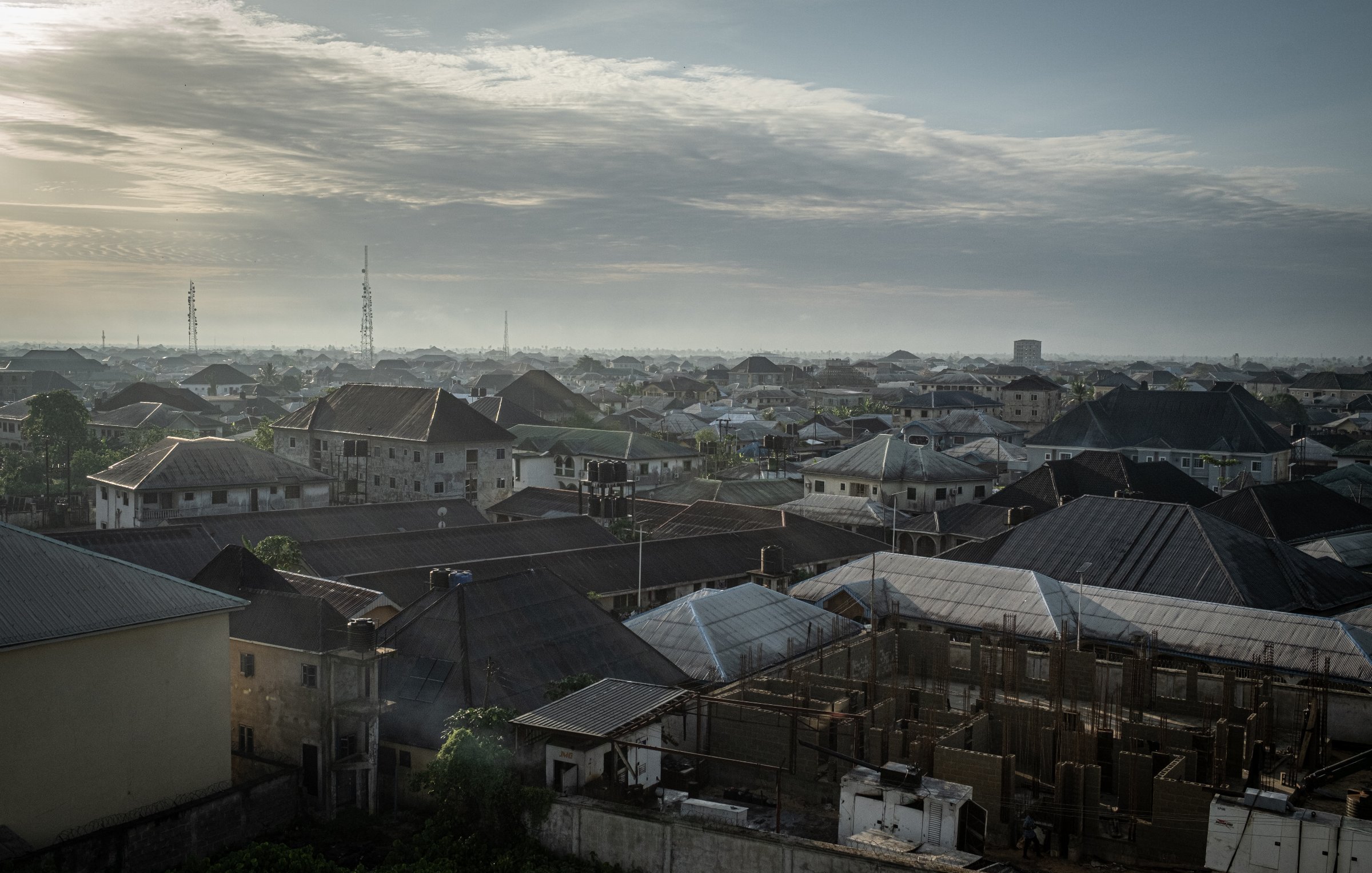 Dans cette image, on voit un paysage urbain d'une ville d'Afrique, probablement au lever du soleil. Les toits de différentes maisons se chevauchent, formant une mosaïque de formes et de textures. La majorité des toits sont en tôle, certains ayant des angles abrupts, tandis que d'autres sont plats. En arrière-plan, on aperçoit des constructions en cours, avec des structures en béton qui indiquent un développement urbain. Le ciel est parsemé de nuages, offrant une lumière douce et diffuse. On devine une atmosphère paisible au matin, avec des ombres légères qui accentuent les contours des bâtiments. Au loin, on distingue des antennes et des colonnes, signes de l'urbanisation moderne. L'ensemble dégage une impression de vie et d'activité, mais aussi de tranquillité à cette heure matinale.