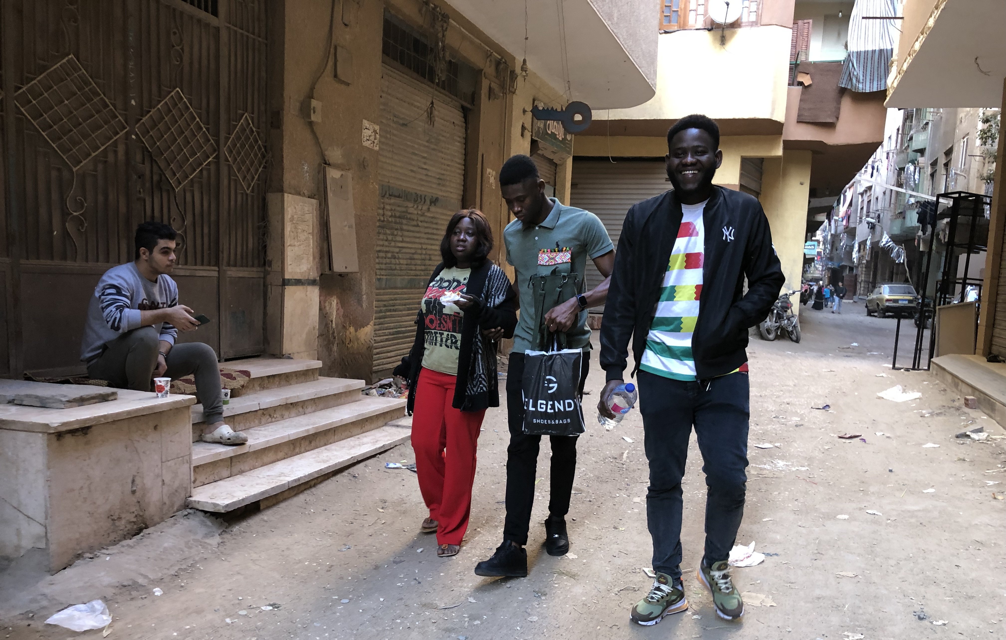 L'image montre une rue étroite et animée dans un quartier urbain. Il y a des déchets dispersés sur le sol, indiquant un environnement un peu désordonné. Sur le côté gauche, un jeune homme est assis sur des escaliers, absorbé par son téléphone ou ses réflexions. Au centre, trois personnes marchent ensemble : une femme portant un haut coloré et un pantalon rouge, un homme en t-shirt gris foncé et un autre homme vêtu d'un haut noir avec des détails colorés. Ils semblent converser joyeusement. En arrière-plan, on peut apercevoir plusieurs bâtiments qui témoignent de la vie locale, avec des balcons et des traces du quotidien. L'atmosphère est dynamique, capturant un moment de vie dans cette rue.