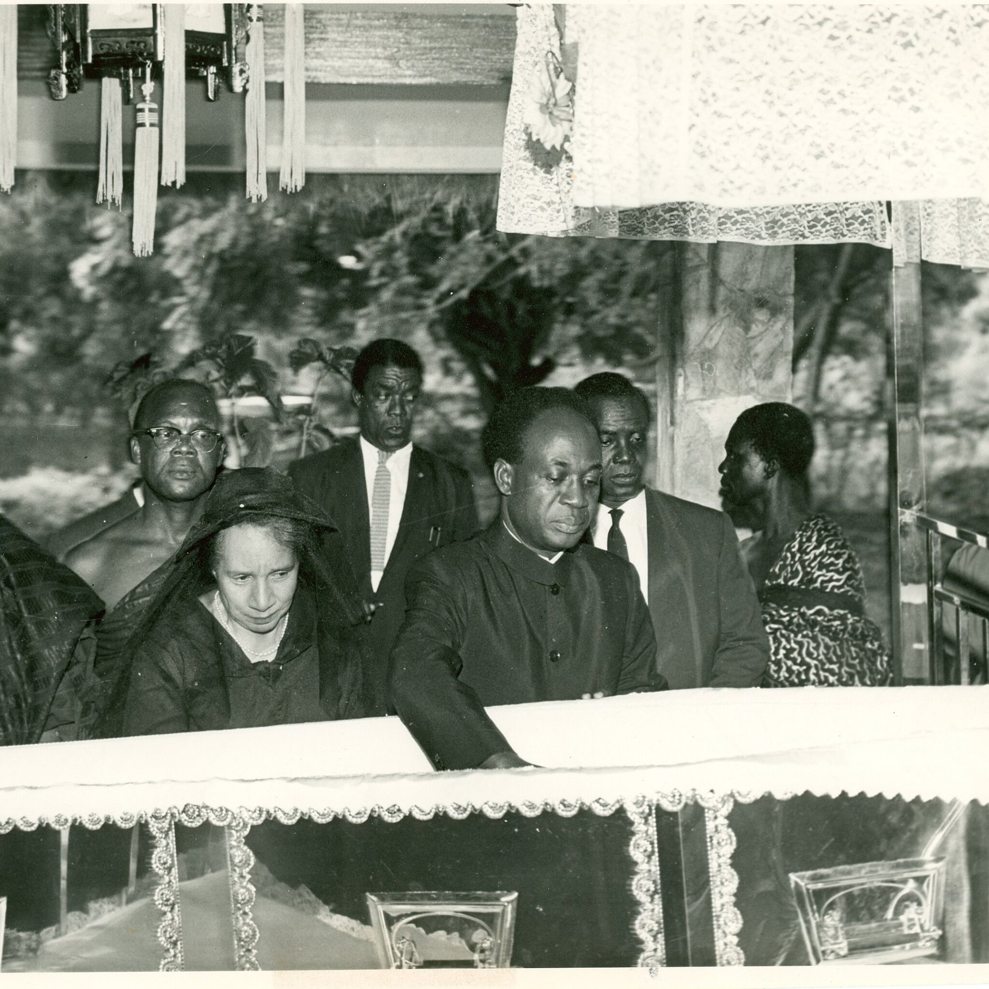 L'image présente une scène de recueillement, probablement lors d'un enterrement ou d'une commémoration. Au premier plan, un homme, habillé en noir, se penche vers un cercueil reposant sur une estrade, symbolisant le respect et la tristesse. À ses côtés se trouve une femme, également en noir, qui semble très émue. En arrière-plan, plusieurs personnes regardent la scène avec un air sérieux, toutes vêtues de manière formelle, ce qui souligne le caractère solennel de l'événement. L'atmosphère est empreinte de respect et de recueillement, avec une ambiance calme, interrompue seulement par les murmures des participants.