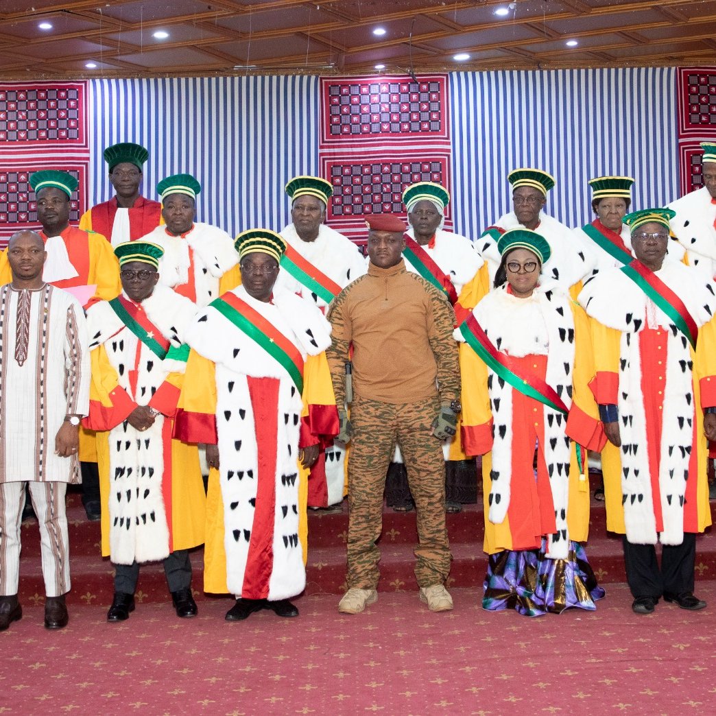 L'image montre un groupe de personnes posant ensemble dans un cadre formel. Au centre, il y a un homme en tenue militaire, entouré par un groupe de personnes habillées en robes hautes avec des motifs distinctifs et des chapeaux colorés. Les vêtements semblent refléter une certaine tradition ou un statut élevé, avec des éléments de couleur rouge, vert et jaune. Les membres du groupe arborent aussi des écharpes qui traversent leur poitrine, ajoutant une touche de distinction. En arrière-plan, il y a des décorations murales et une scène de cérémonie qui donne une atmosphère solennelle à l'ensemble. Le sol est recouvert d'un tapis rouge, contribuant à l'élégance du moment.