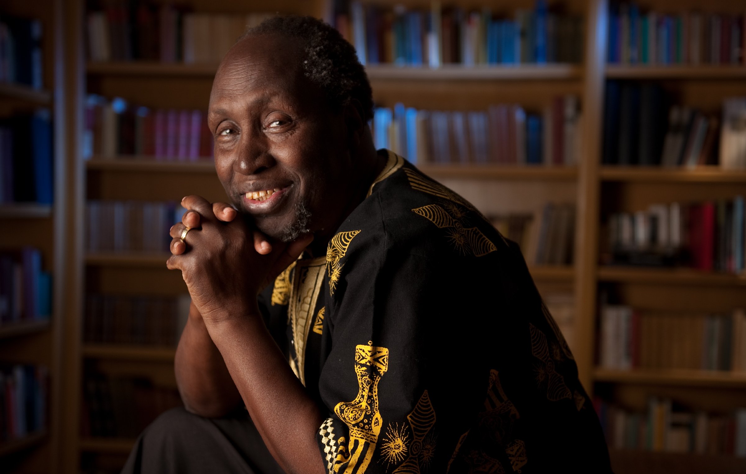 L'image montre un homme assis dans une bibliothèque, entouré de nombreuses étagères remplies de livres. Il a un sourire chaleureux et une expression amicale. Il porte une chemise noire ornée de motifs dorés, ce qui ajoute une touche d'élégance à son apparence. Son visage exprime une sagesse et une bienveillance, tandis qu'il a les mains croisées sous son menton. L'arrière-plan est légèrement flou, mettant en valeur sa figure et créant une atmosphère accueillante et studieuse.