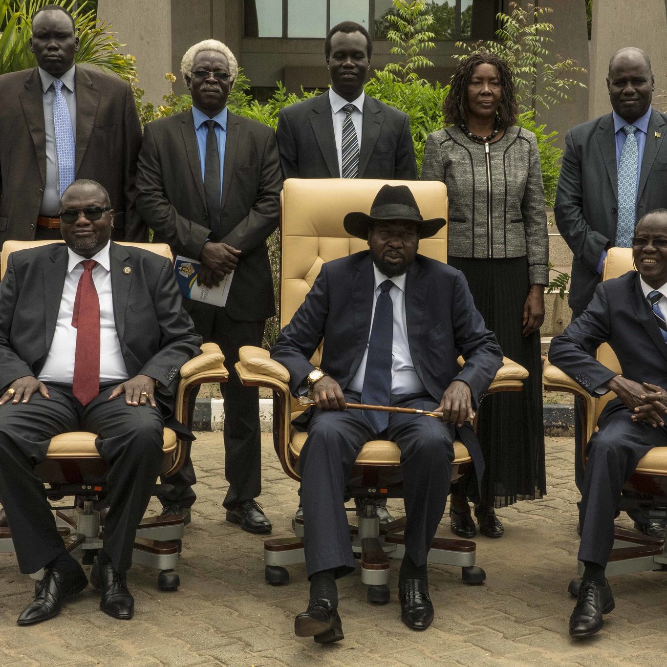 L'image présente un groupe de personnes posant ensemble à l'extérieur d'un bâtiment. Au centre, on voit trois hommes assis dans de grandes chaises confortables. Le premier homme à gauche porte des lunettes, un costume sombre et une cravate rouge. Le second, au centre, est vêtu d'un costume noir, d'une cravate et d'un chapeau noir, et semble être le leader du groupe. Le troisième homme, à droite, porte également un costume et une cravate. Derrière eux, plusieurs personnes se tiennent debout, toutes en tenue formelle. L'environnement est verdoyant, avec des plantes et un chemin en pavé. L'atmosphère semble sérieuse et officielle, suggérant une rencontre importante.