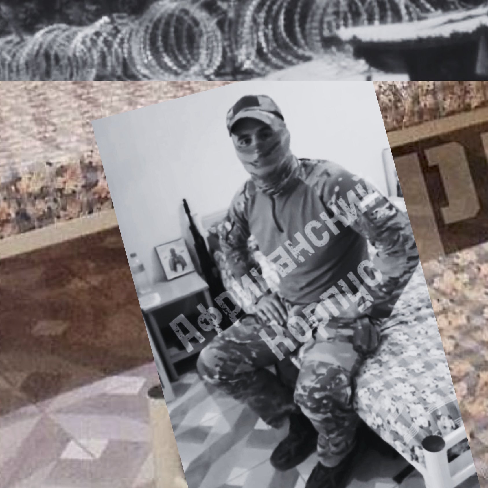 L'image montre un intérieur, probablement d'une installation militaire ou d'un camp. Il y a un lit avec un matelas couvert d'un drap à motifs floraux, et à côté se trouve une arme à feu posée sur le sol. Un homme, vêtu d'un uniforme militaire et portant un masque, est assis sur le lit. Des barbelés sont visibles à l'arrière-plan, ce qui suggère un environnement fortifié. Le texte en cyrillique sur la photo semble indiquer un message ou un titre, mais je ne peux pas le traduire. L'atmosphère générale semble sérieuse et militaire.